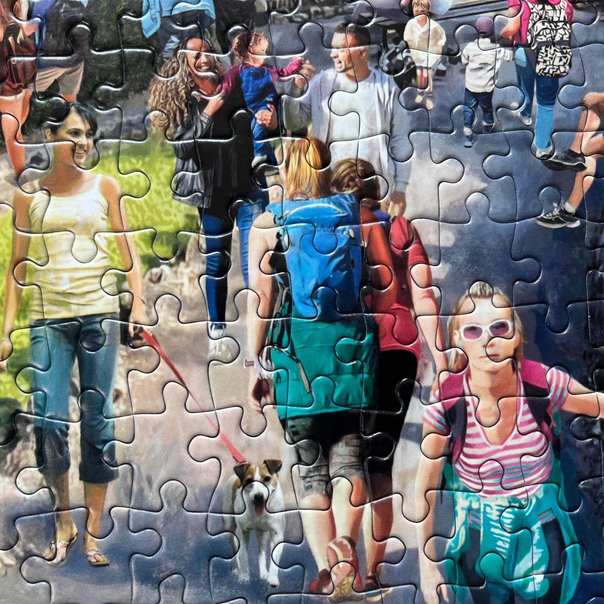 A completed jigsaw puzzle showing a colourful collage of people walking dogs and strolling outdoors on a path, with puzzle pieces still visible around the edges.
