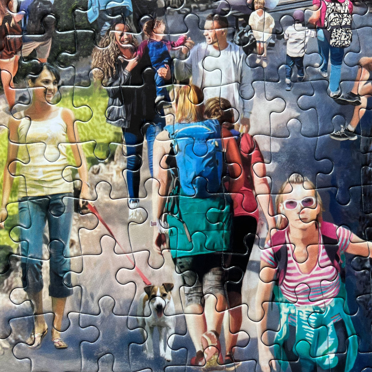 A completed jigsaw puzzle showing a colourful collage of people walking dogs and strolling outdoors on a path, with puzzle pieces still visible around the edges.