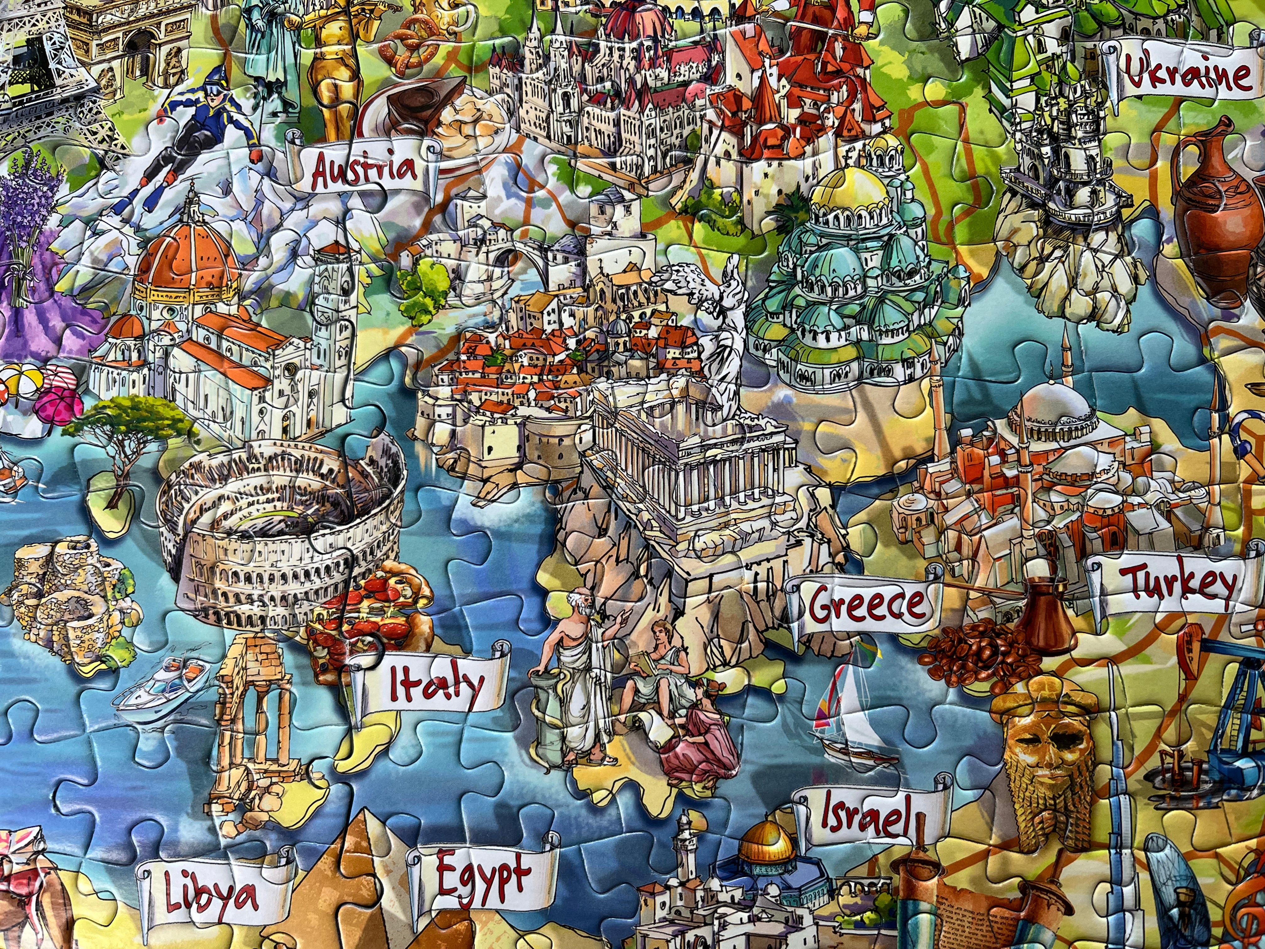 A colourful illustrated jigsaw puzzle showing a map-style collage of Mediterranean and Eastern European countries, with labels such as Austria, Italy, Greece, Turkey, Israel, Libya, and Ukraine. The puzzle pieces are assembled and the artwork fills the frame.