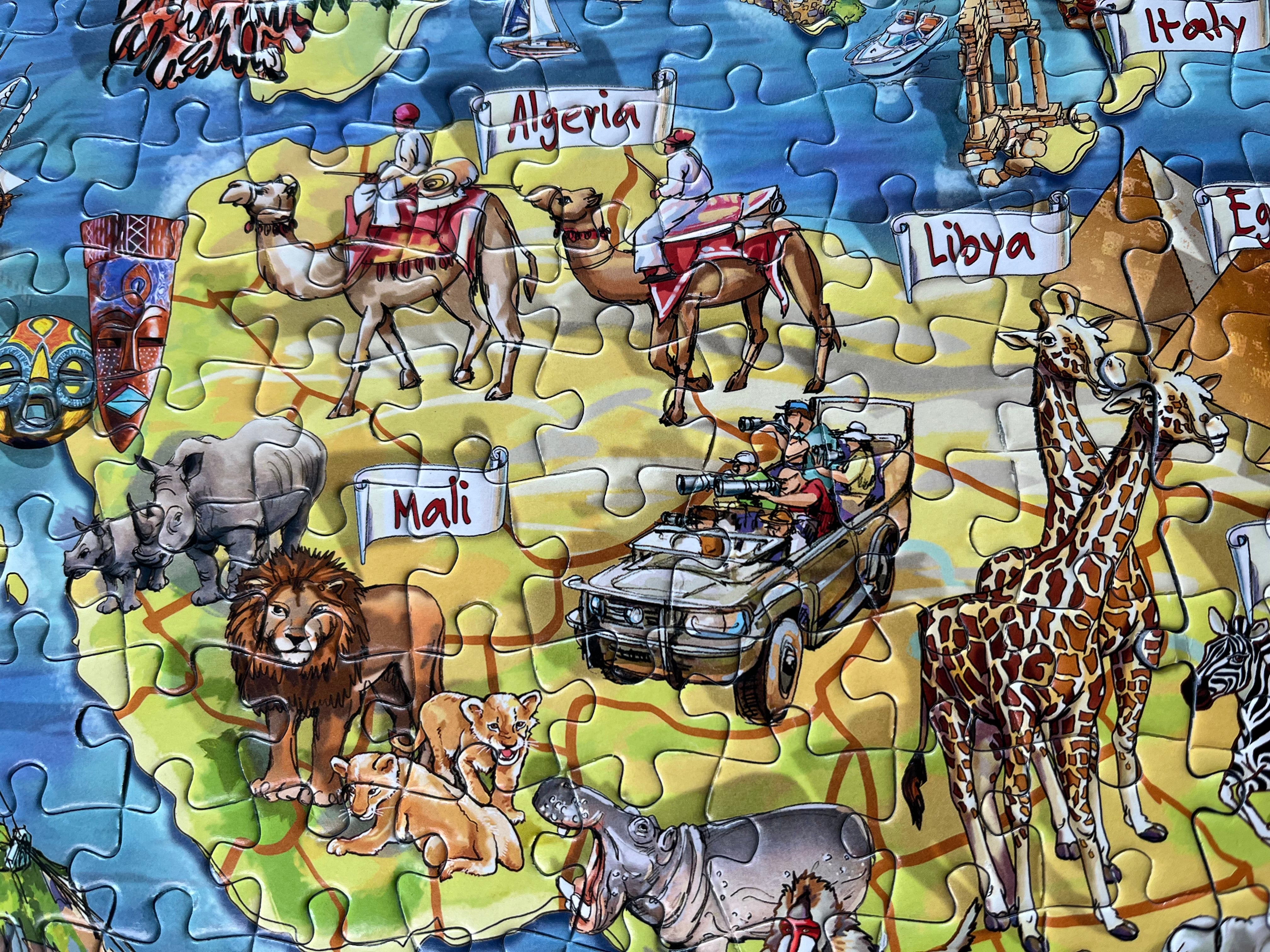 A colourful jigsaw puzzle depicting a map of Africa with labelled countries such as Algeria, Libya, and Mali, plus illustrated animals including camels, lions, giraffes, rhinos, zebras, and a safari vehicle.