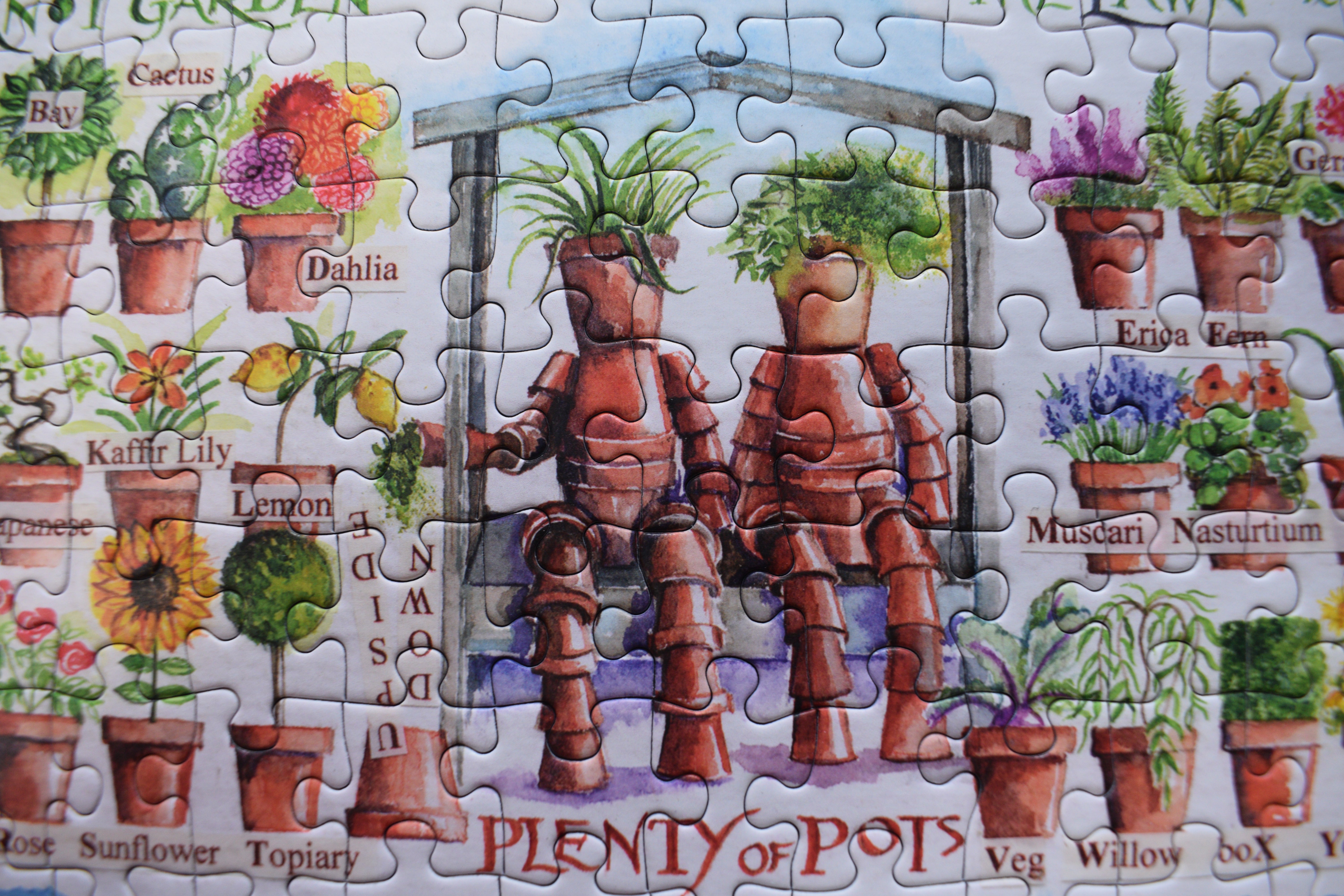 A completed jigsaw puzzle featuring a whimsical garden scene with two terracotta planter robots sitting in front of a greenhouse, surrounded by labeled illustrated potted plants such as Cactus, Dahlia, Lemon, and Sunflower on a white puzzle background.