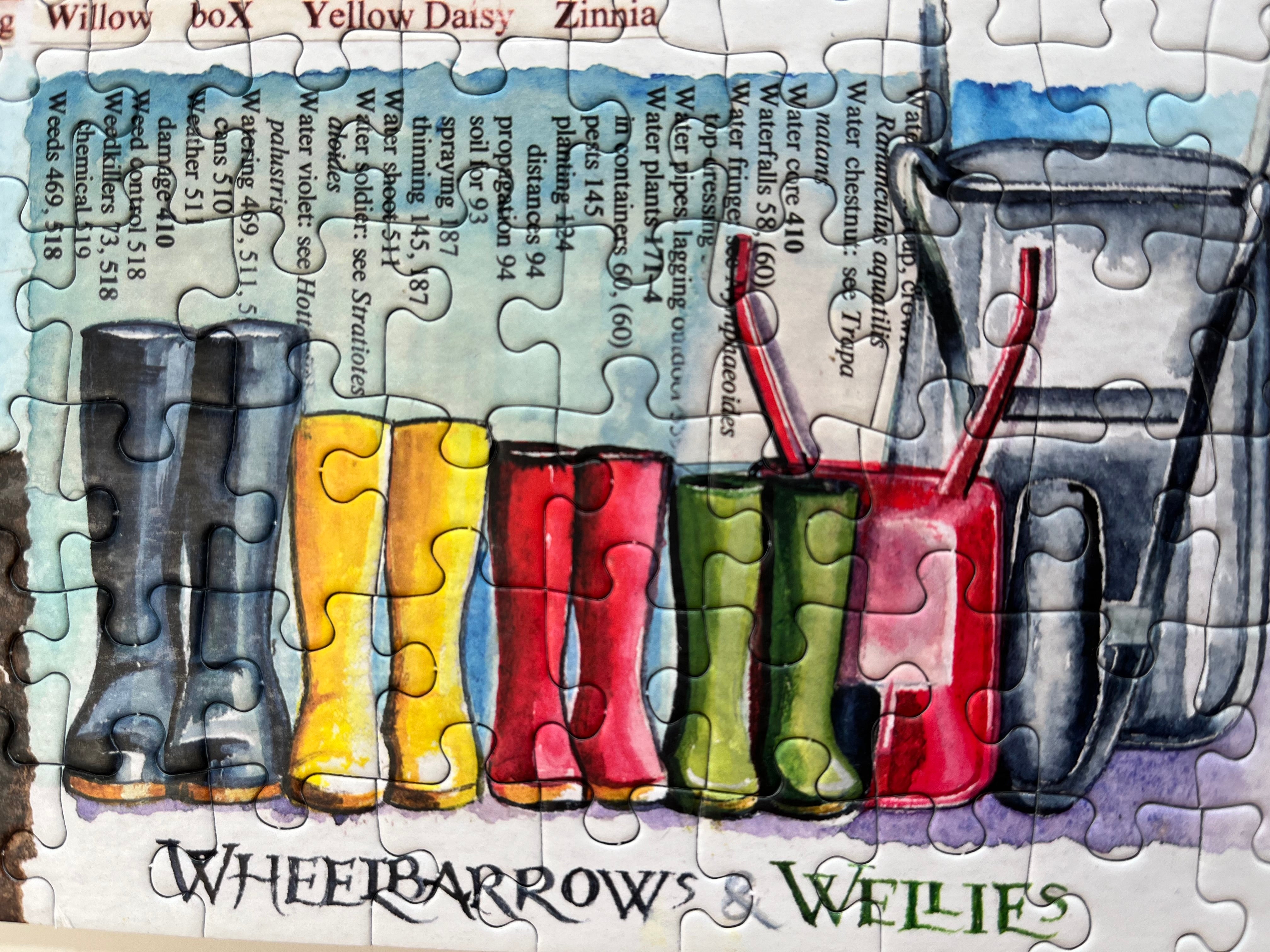 A close-up of a jigsaw puzzle featuring a watercolour-style illustration of colourful rain boots and a watering can, with the text 'WHEELBARROWS & WELLIES' at the bottom.