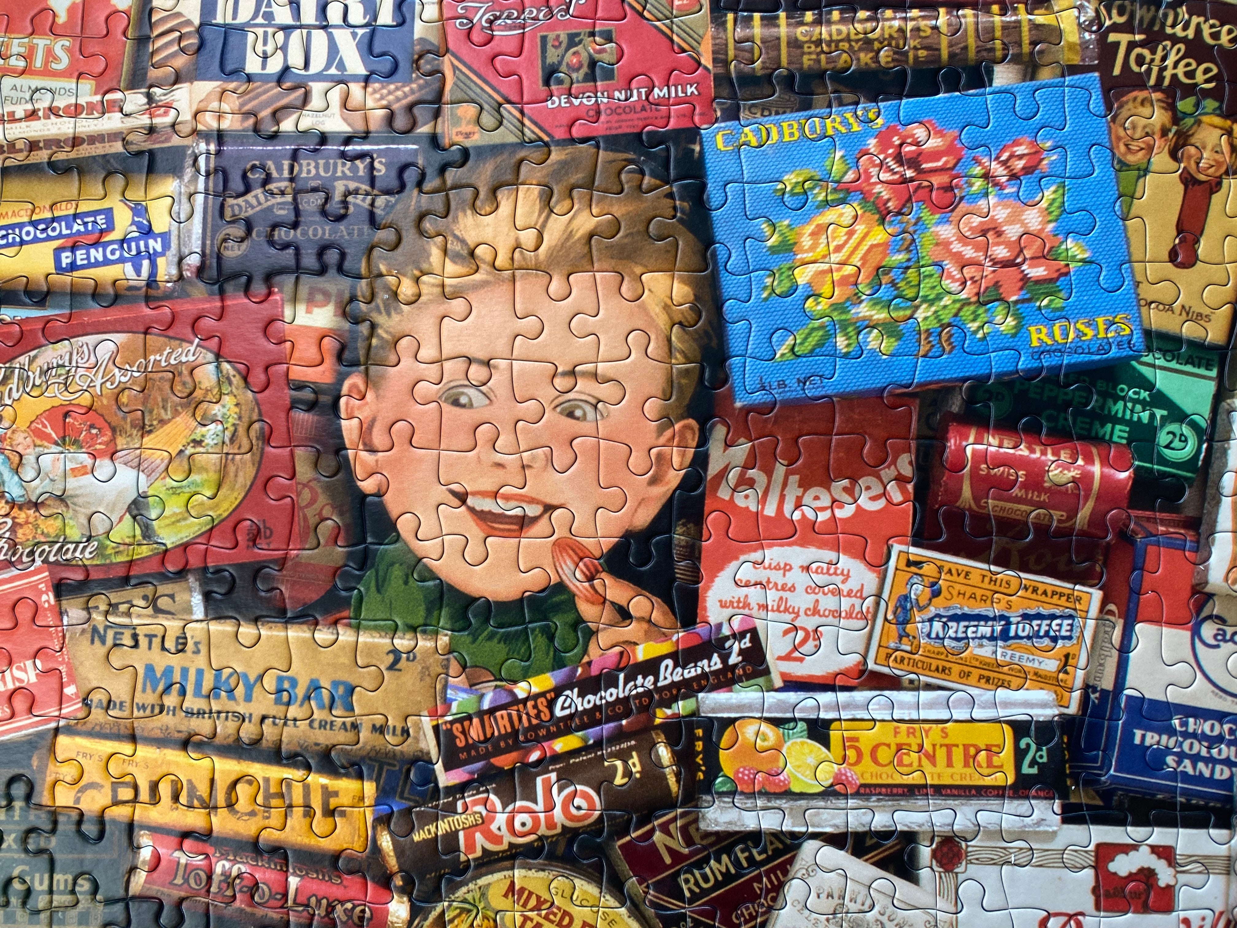 A completed jigsaw puzzle featuring a collage of vintage candy and chocolate boxes with a large illustrated child's face in the center, photographed straight-on against no separate background because the puzzle fills the frame.