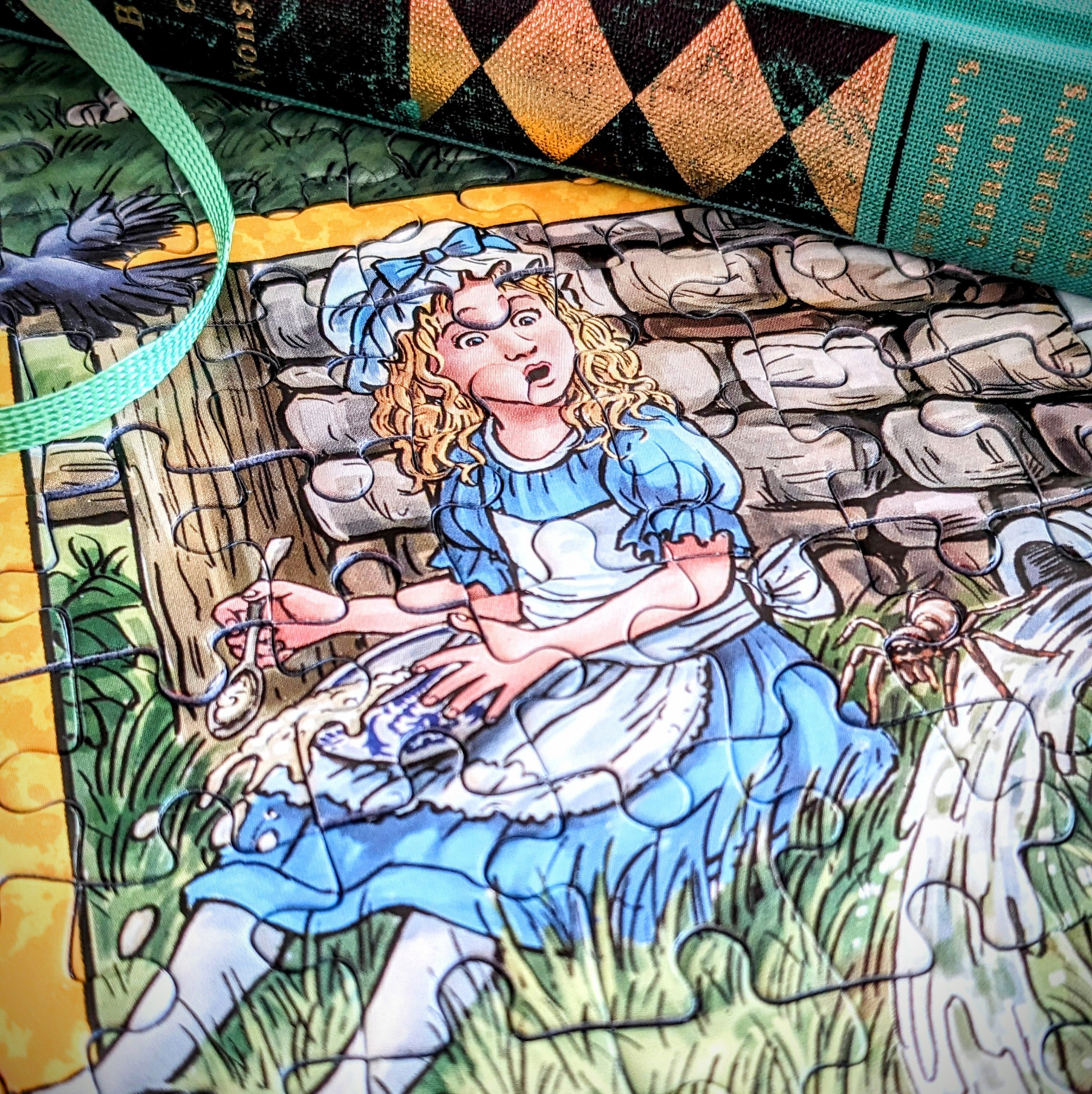 A close-up of a printed fabric surface featuring an illustrated scene of Alice in a blue dress sitting in grass in front of a stone wall, with part of a green bag strap and a patterned green-and-black item visible around the edges.