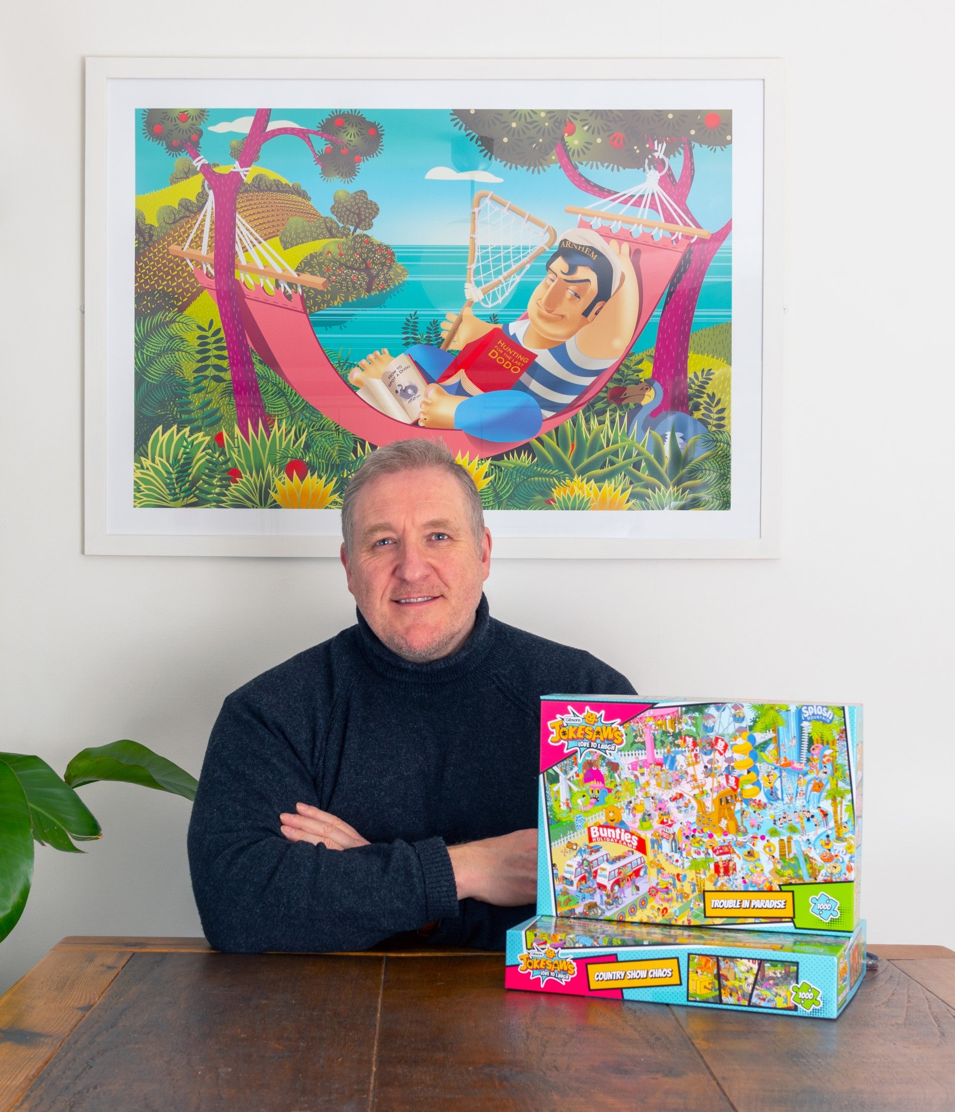 Man sitting at a table with a colorful board game box, framed artwork on the wall.
