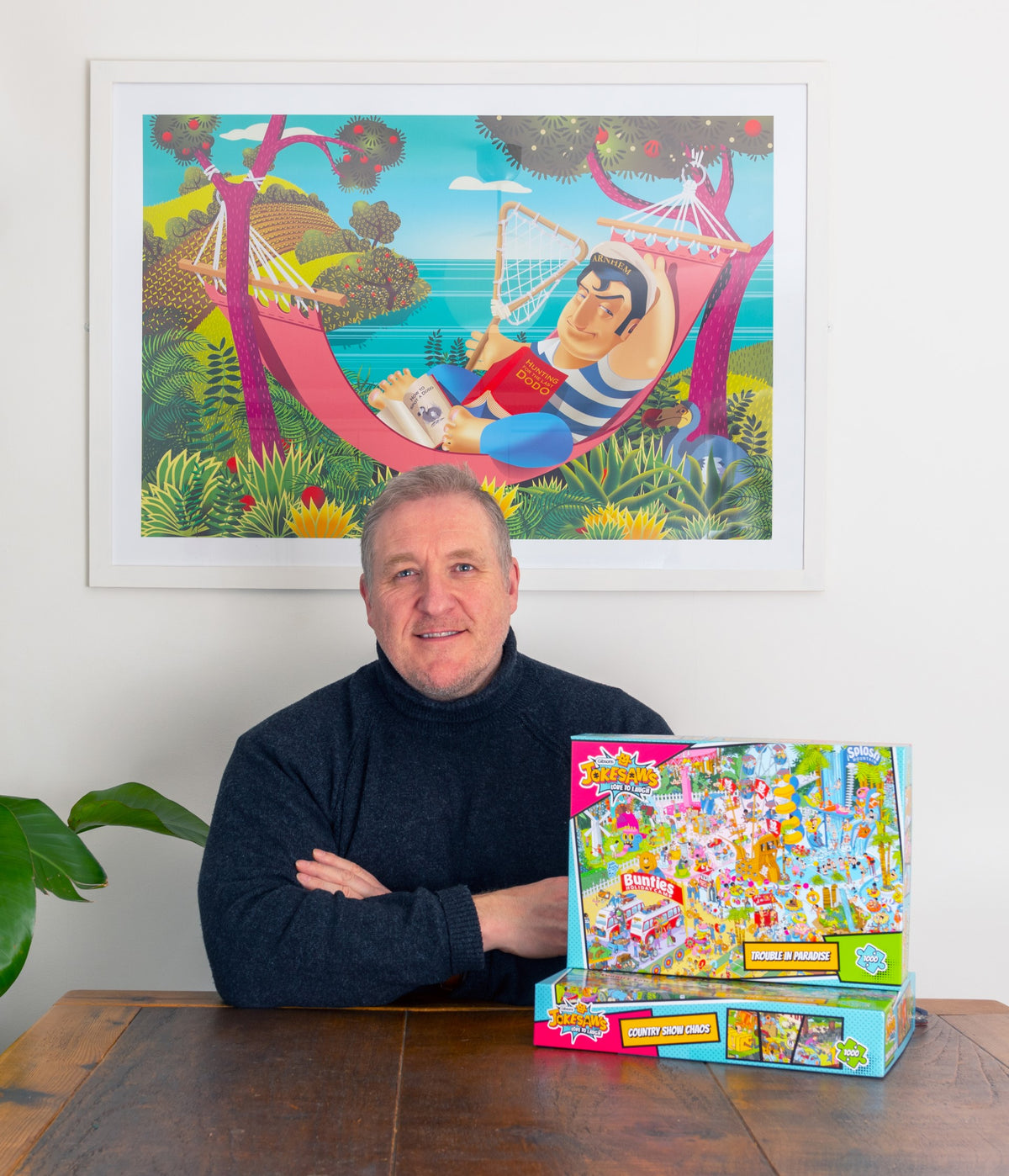 Man sitting at a table with a colorful board game box, framed artwork on the wall.