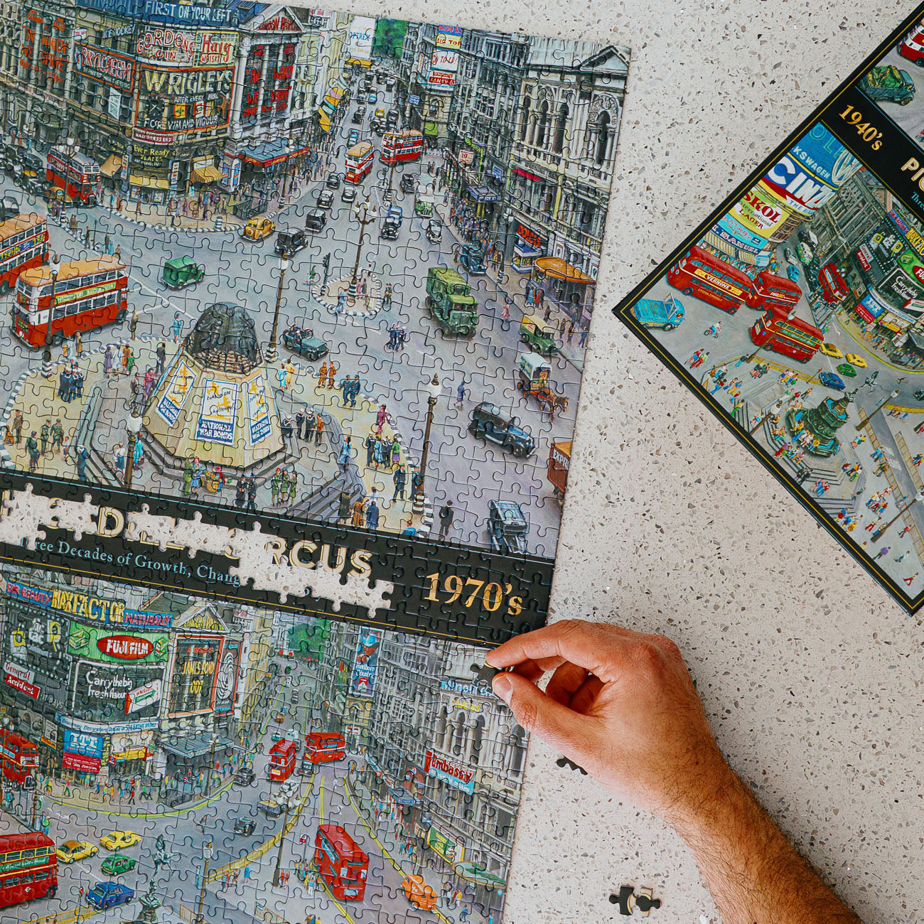 A hand is assembling a large jigsaw puzzle depicting a busy 1970s London street scene with red buses, traffic, and storefronts. The puzzle is shown partly completed on a speckled white surface, with the puzzle box visible in the upper right corner.