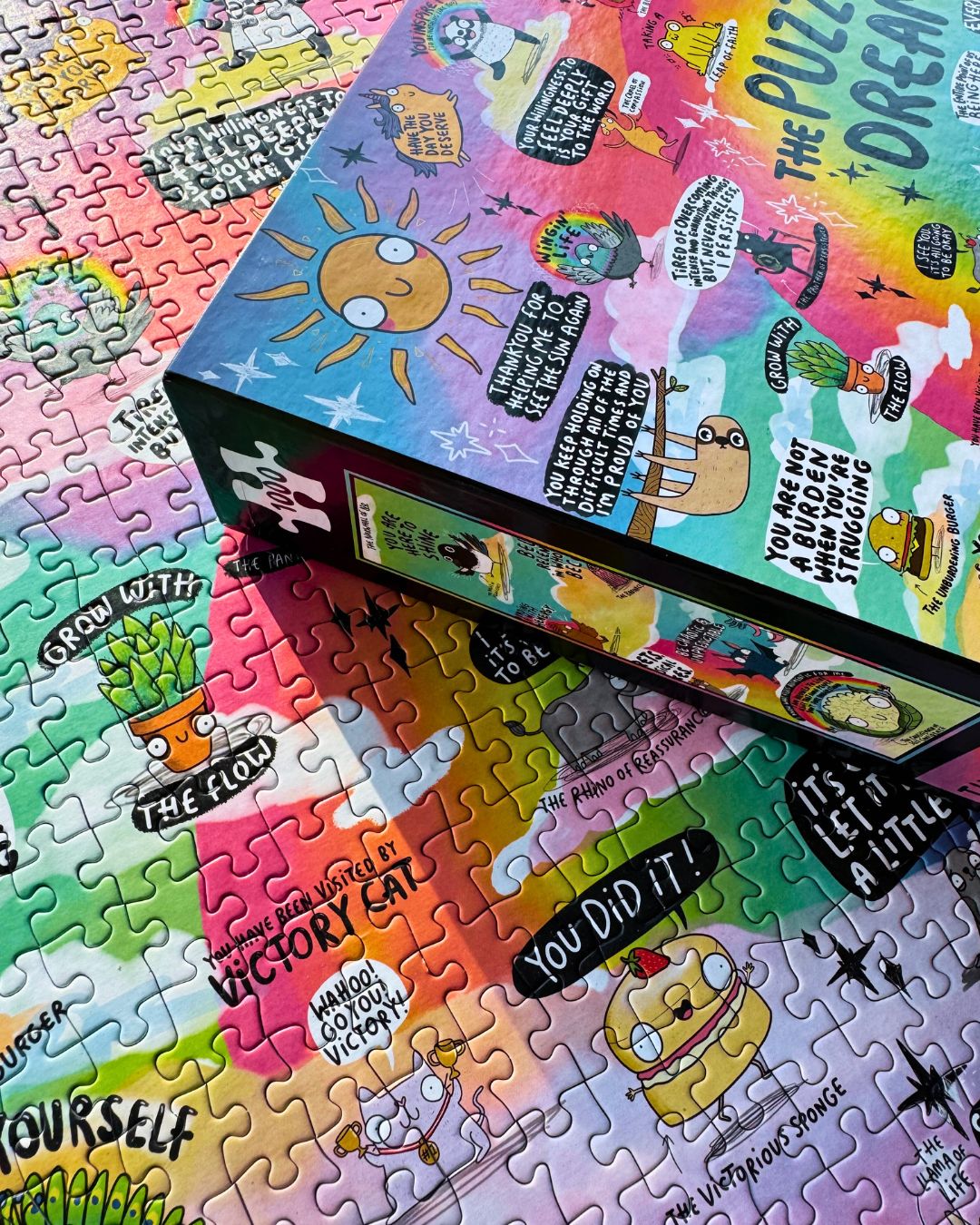 A colourful jigsaw puzzle box with cartoon illustrations and motivational quotes, placed diagonally on top of a completed multicolour jigsaw puzzle.