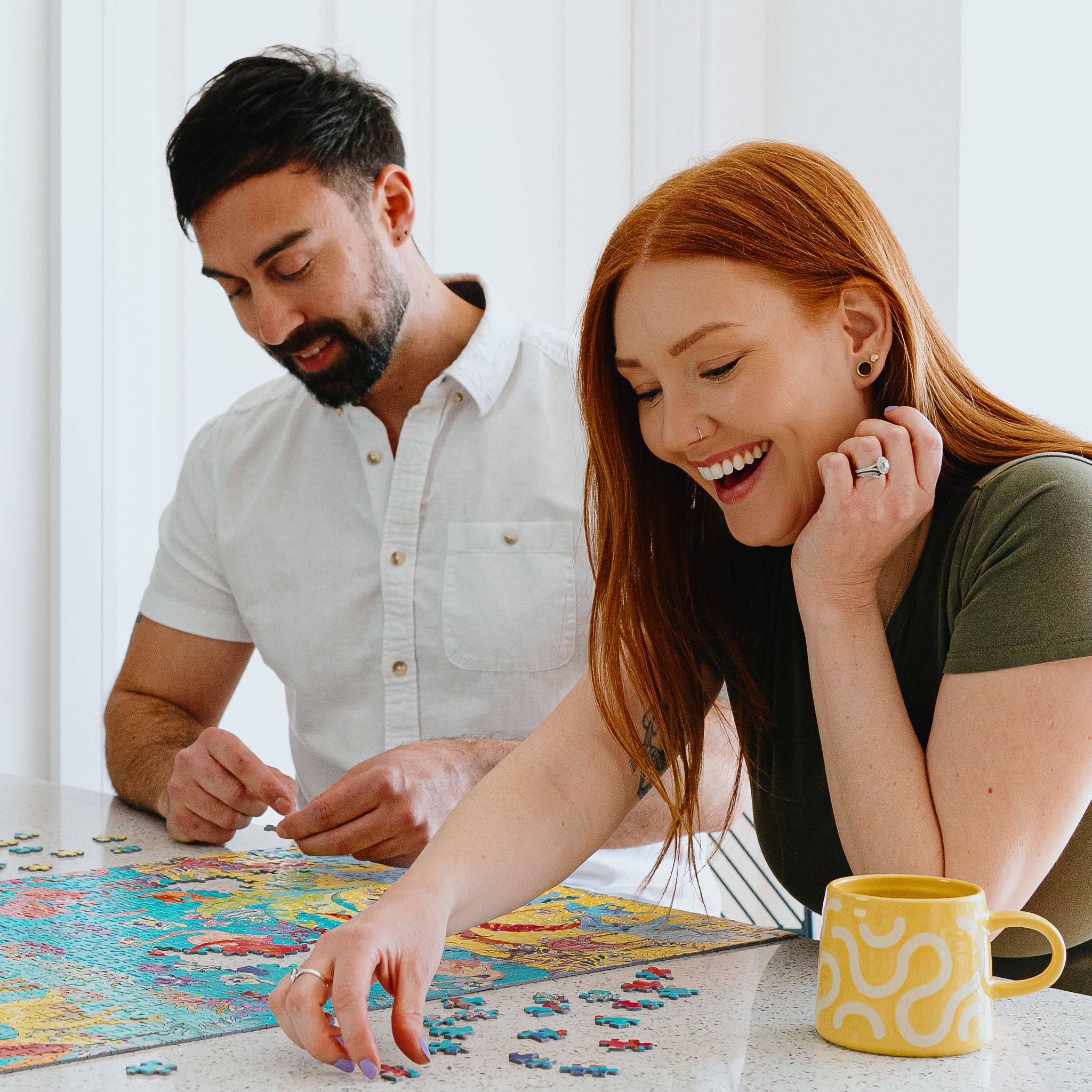 A man and a woman laughing while playing a board game at a table, with a yellow patterned mug on the counter in a bright indoor setting.