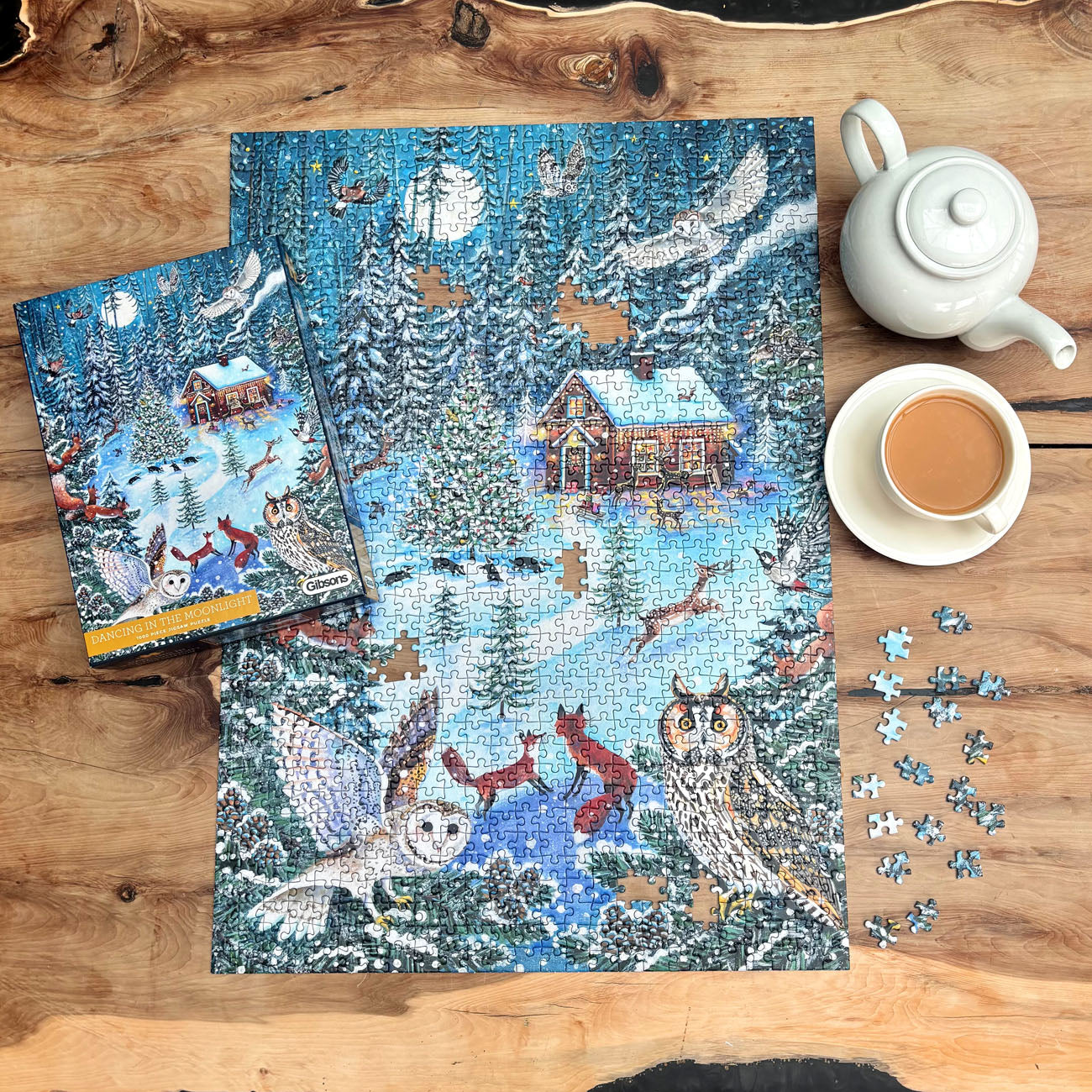 A completed winter forest jigsaw puzzle with its matching box is displayed on a rustic wooden table, alongside a white teapot, a cup of coffee, and a few loose puzzle pieces. The puzzle features a cozy cabin, snowy trees, owls, foxes, and other winter animals under a full moon.