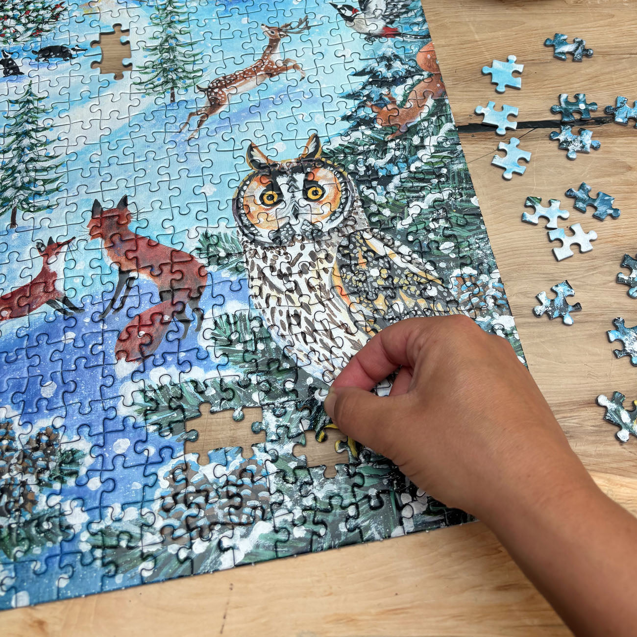 A person's hand placing a jigsaw puzzle piece onto a completed winter forest puzzle featuring an owl, fox, deer, and snowy trees, on a light wooden table with loose puzzle pieces scattered nearby.