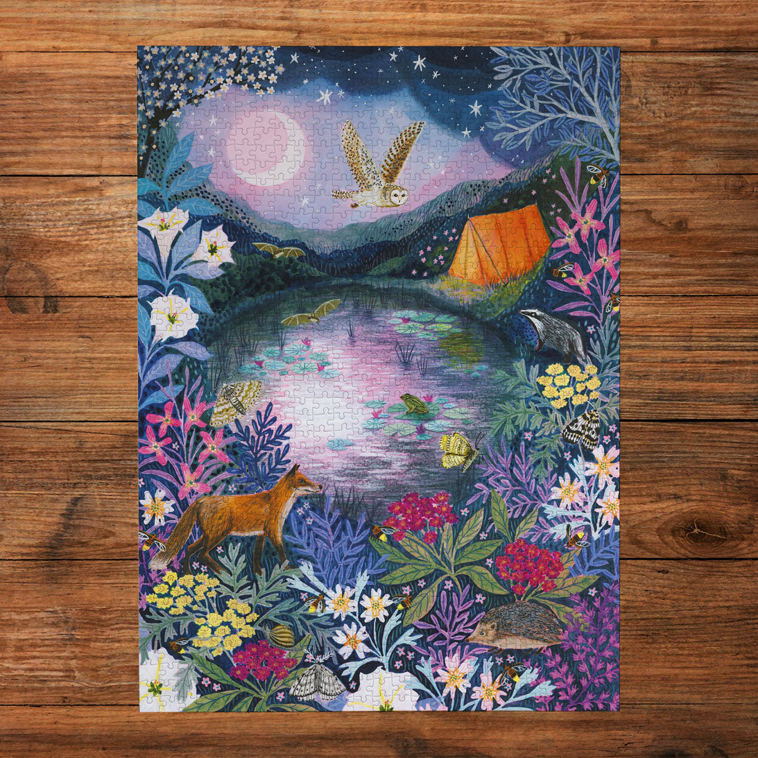 An art puzzle featuring a whimsical woodland night scene, displayed flat on a rustic brown wooden surface. The artwork depicts an owl flying over a moonlit pond with a glowing orange tent, surrounded by colourful wildflowers, a fox, a badger, and a hedgehog under a starry sky.