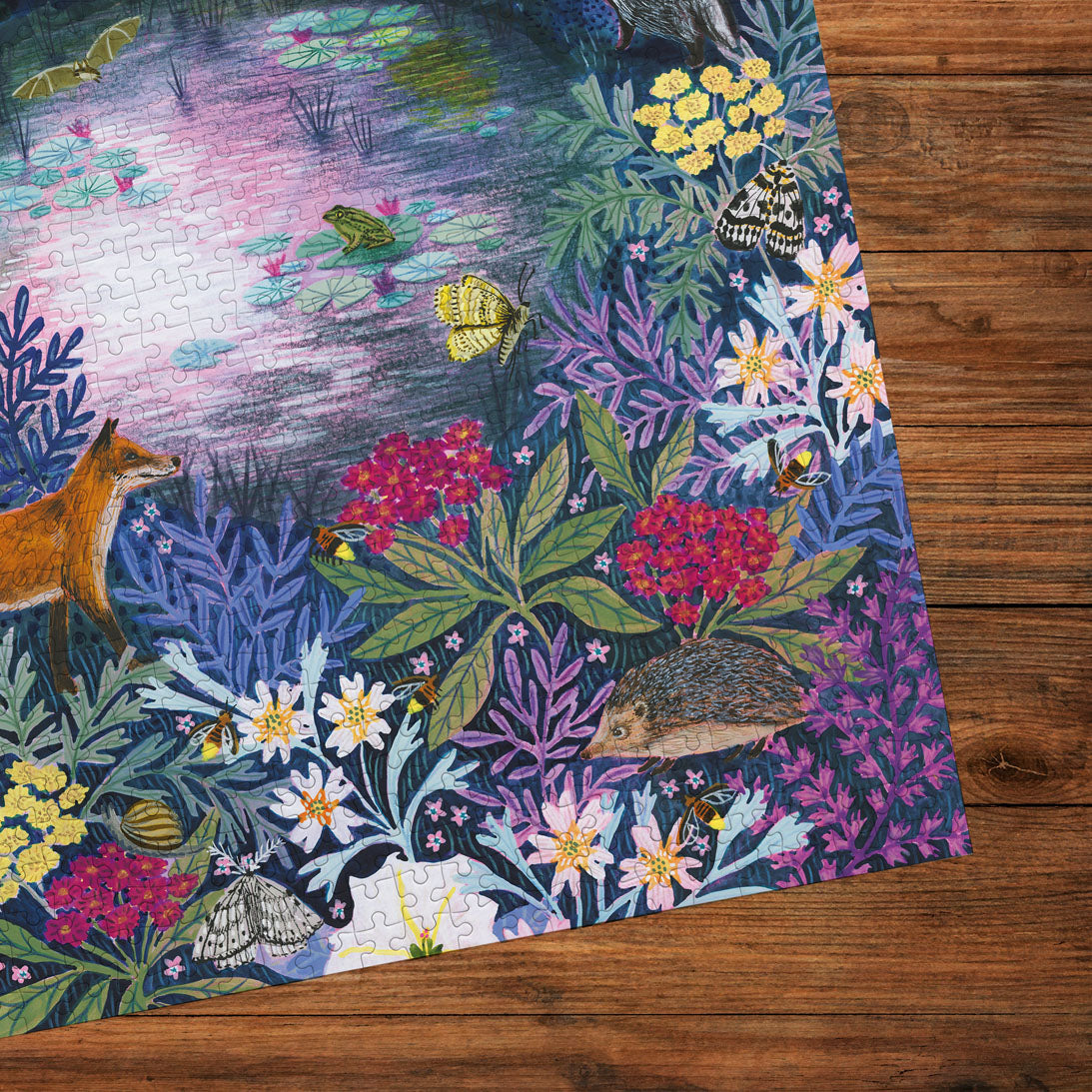 A completed jigsaw puzzle featuring a detailed woodland scene with a fox, hedgehog, frog, butterflies, flowers, and birds, placed on a dark wood surface.
