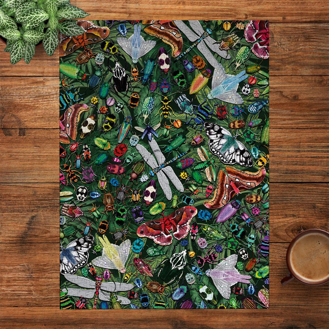 A rectangular art print featuring a dense, colourful pattern of dragonflies, butterflies, moths, bees, skulls, and small insects on a dark green background, styled flat on a wooden tabletop with a plant leaf in the top-left corner and a cup of coffee in the bottom-right corner.