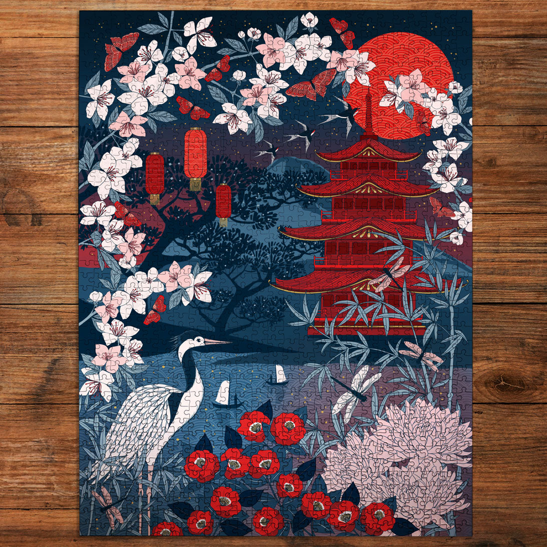 A rectangular art jigsaw puzzle laid flat on a wooden surface, featuring a stylized night scene with a red pagoda, red moon, pink blossoms, a white crane, red flowers, and dark blue foliage.