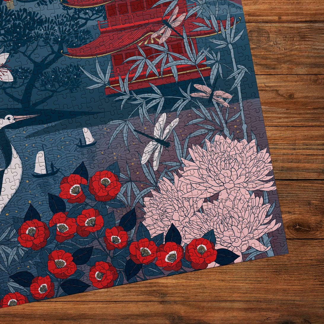 A close-up of a completed jigsaw puzzle featuring a stylized East Asian-inspired scene with red flowers, pink chrysanthemums, cranes, bamboo, a pagoda, and dragonflies on a dark blue background, placed on a wooden surface.