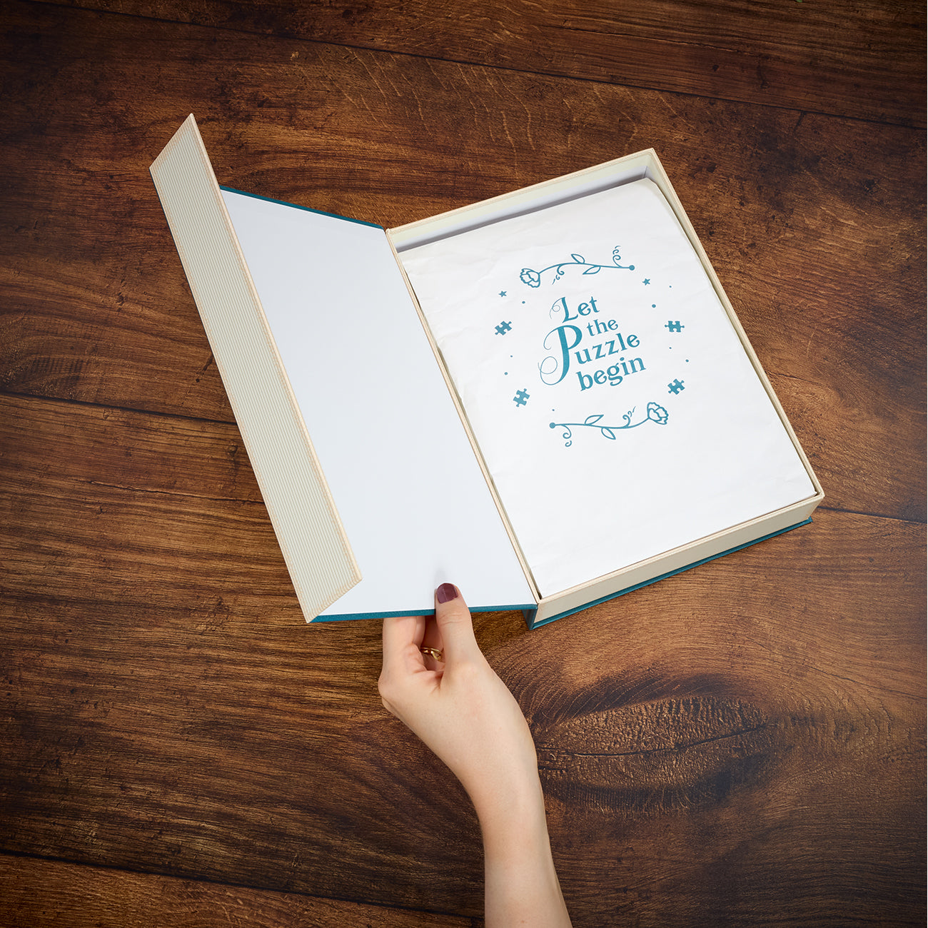 An open cream-colored gift box with a blue inner border sits on a dark wooden table. Inside, a white envelope or card is held open by a person's hand, showing the printed phrase 'Let the Puzzle begin' in blue decorative script.