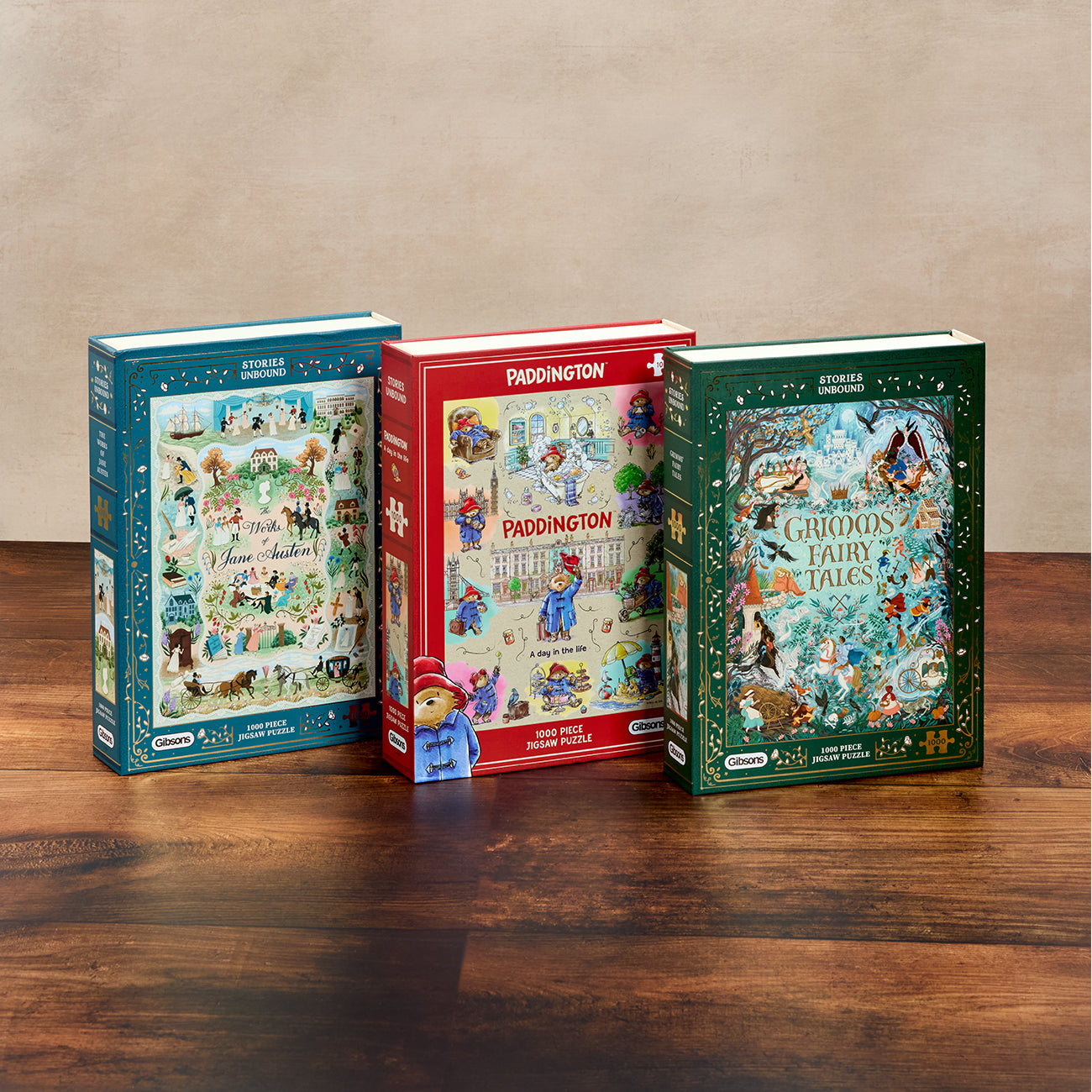 Three boxed children's jigsaw puzzles stand on a wooden tabletop against a beige wall. The left box is blue, the centre box is red and branded Paddington, with a small Paddington figure in front; the right box is green and titled 'Grimm's Fairy Tales'.