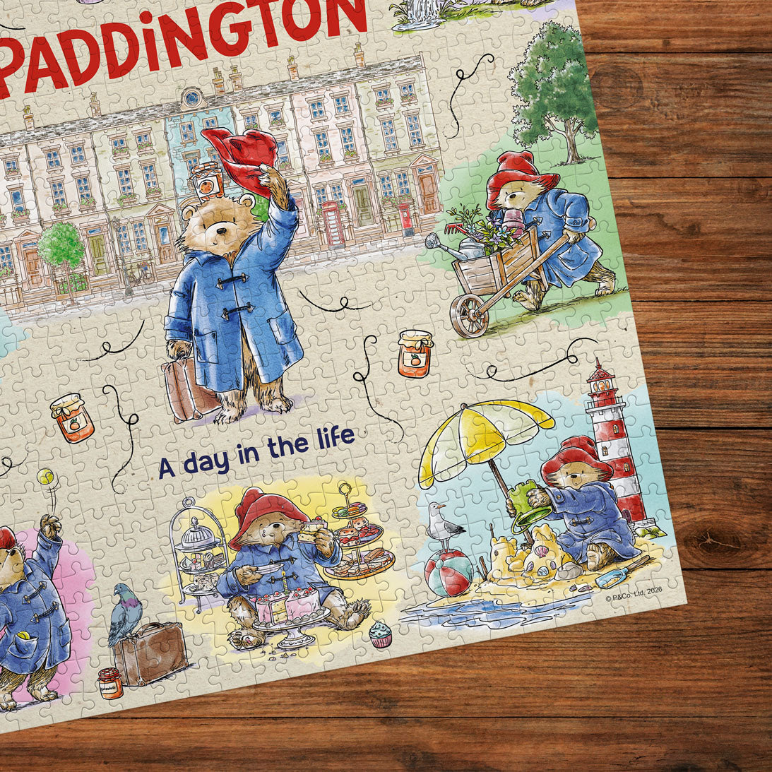 A completed jigsaw puzzle featuring Paddington Bear illustrations and the title 'A day in the life' on a wooden tabletop. The puzzle shows Paddington in blue coats engaging in various activities, with the word 'PADDINGTON' partially visible at the top.