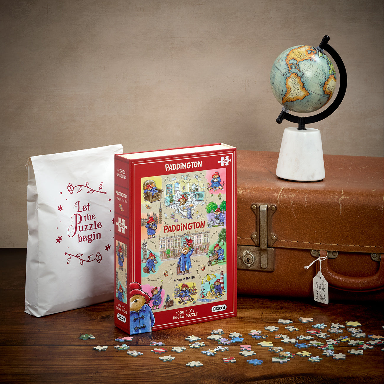 Paddington-themed jigsaw puzzle box displayed on a wooden surface with scattered puzzle pieces in front, a white paper bag reading 'Let the puzzle begin,' and a vintage brown suitcase with a globe on top, all against a neutral studio backdrop.