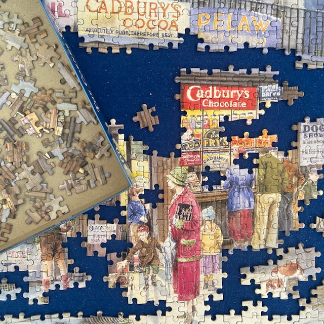 A completed jigsaw puzzle showing a vintage street scene with people and multiple Cadbury's logos, alongside an open box of loose puzzle pieces on a light wood surface.