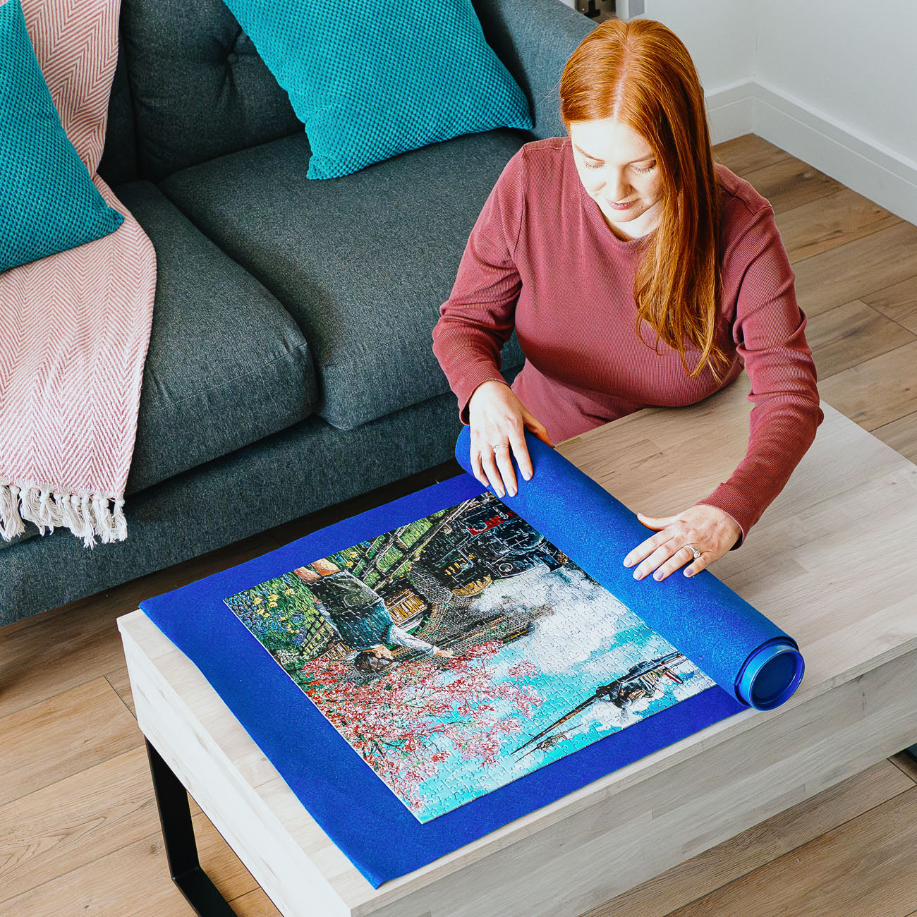 A woman with red hair sits by a gray sofa in a living room, unrolling a blue puzzle mat on a light wood table. The puzzle features a colourful scene of a boy  with a pink blossoms tree and passing by steam train.