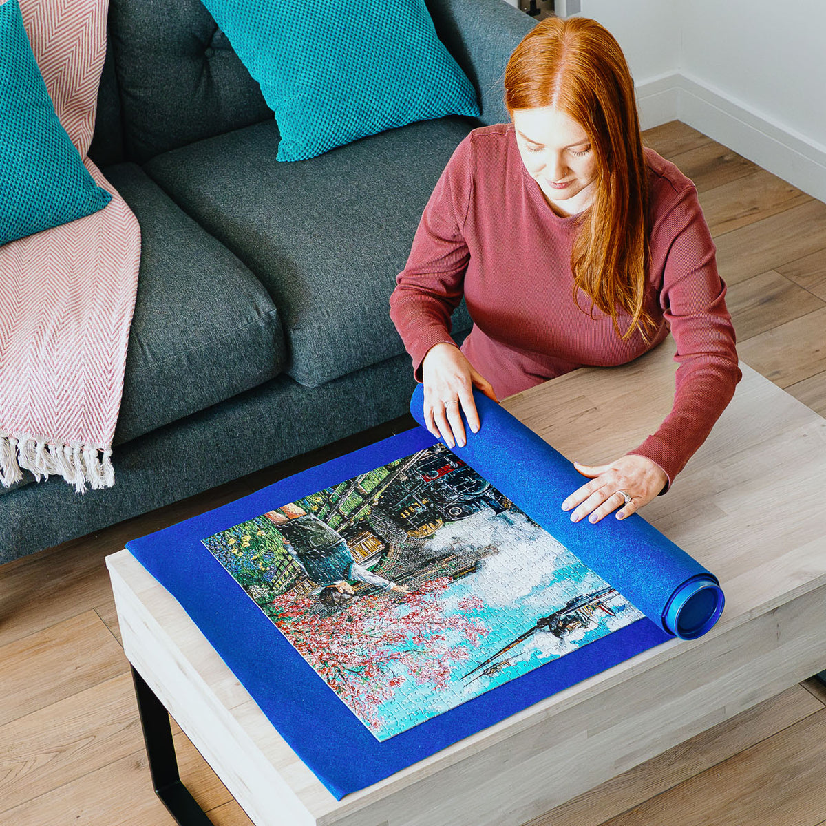 A woman with red hair sits by a gray sofa in a living room, unrolling a blue puzzle mat on a light wood table. The puzzle features a colourful scene of a boy  with a pink blossoms tree and passing by steam train.
