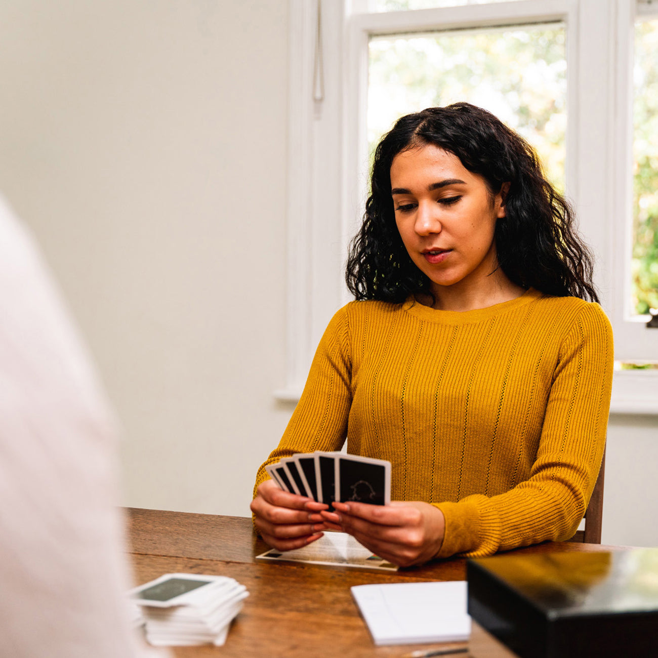 A woman with dark curly hair wearing a mustard yellow ribbed sweater sits at a wooden table, holding a fan of playing cards. A box, a deck of cards, and a notepad are on the table in a brightly lit room.