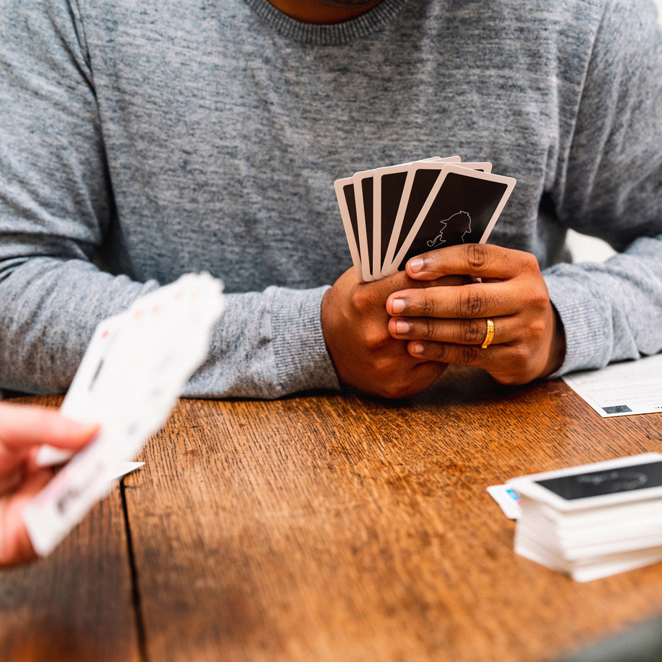 A person in a gray long-sleeve shirt holds a fan of playing cards over a wooden table, with another hand holding cards in the foreground and a small stack of cards on the table.