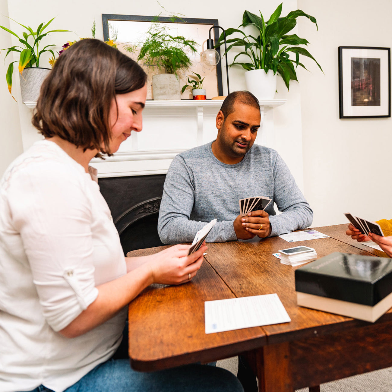 Three people sitting around a wooden table playing a card game in a cozy room with potted plants and framed wall art in the background.