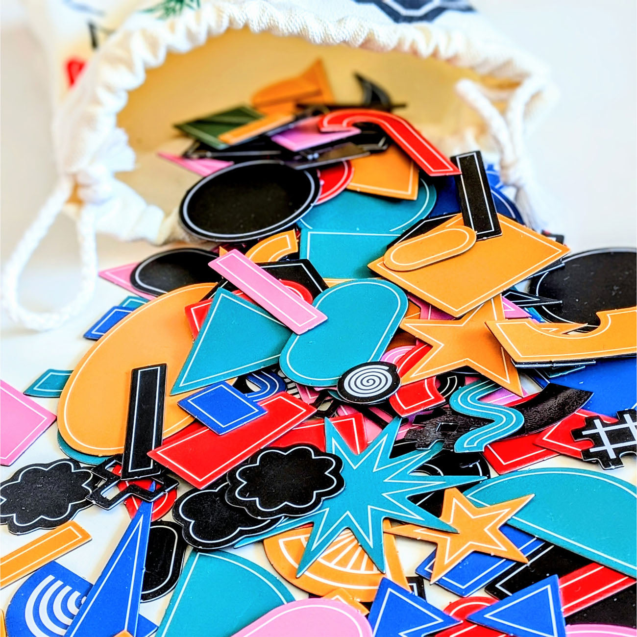 A pile of colourful, flat decorative magnetic stickers or craft pieces in various shapes like stars, clouds, and geometric patterns, spilling out of a white drawstring bag.