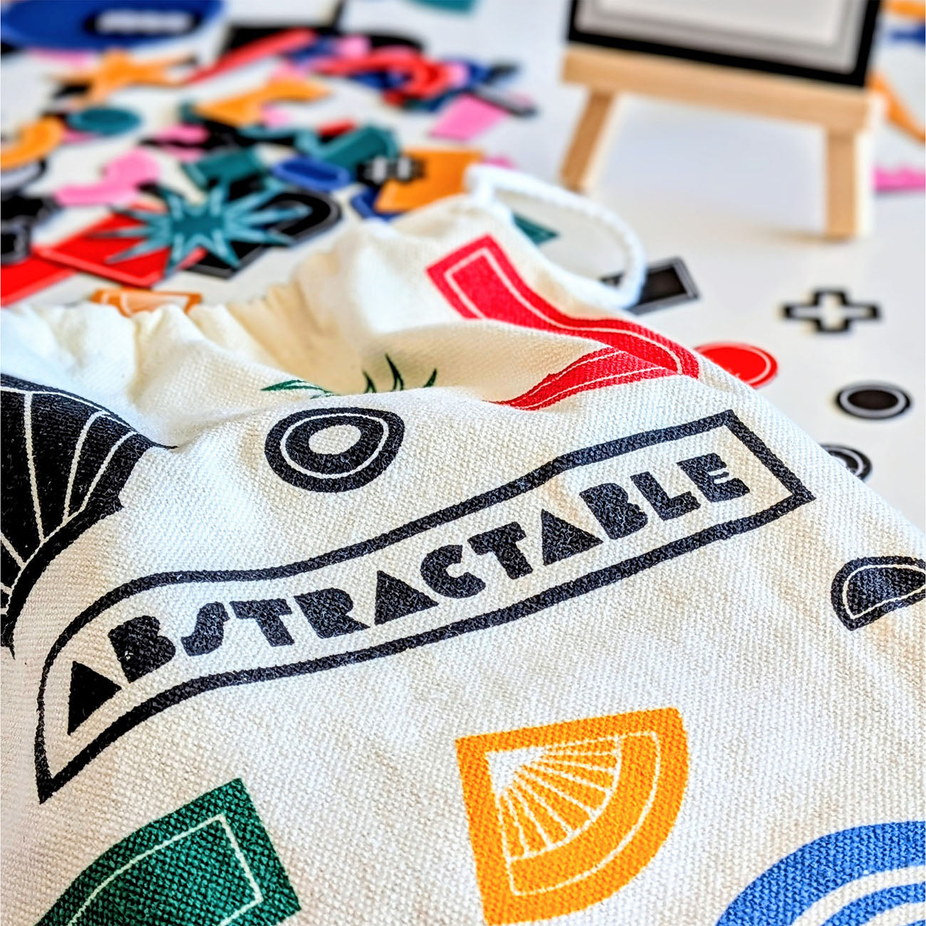A close-up of a cream canvas drawstring bag printed with the word 'ABSTRACTABLE' in black inside a banner, surrounded by colourful abstract geometric shapes. The bag is shown on a white surface with a small wooden easel and more colourful pieces in the background.