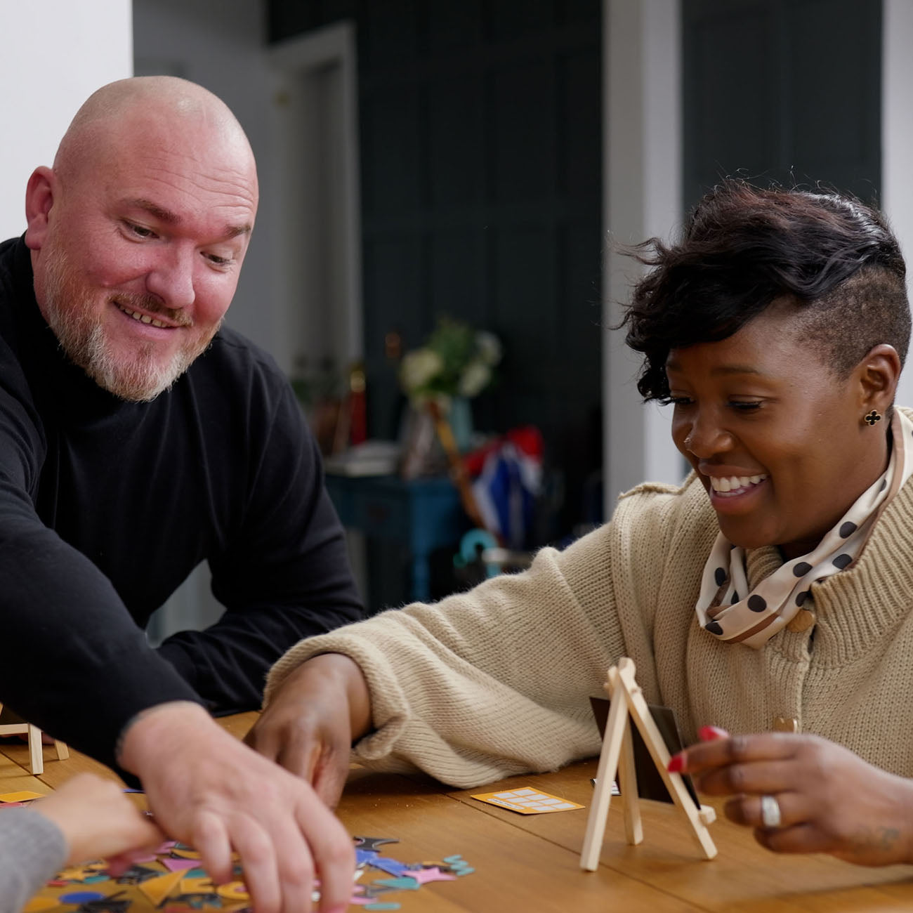 A smiling man and woman sitting together at a wooden table, playing a fun game. The man wears a black long-sleeve shirt, and the woman wears a beige knit sweater and a beige scarf with black polka dots.
