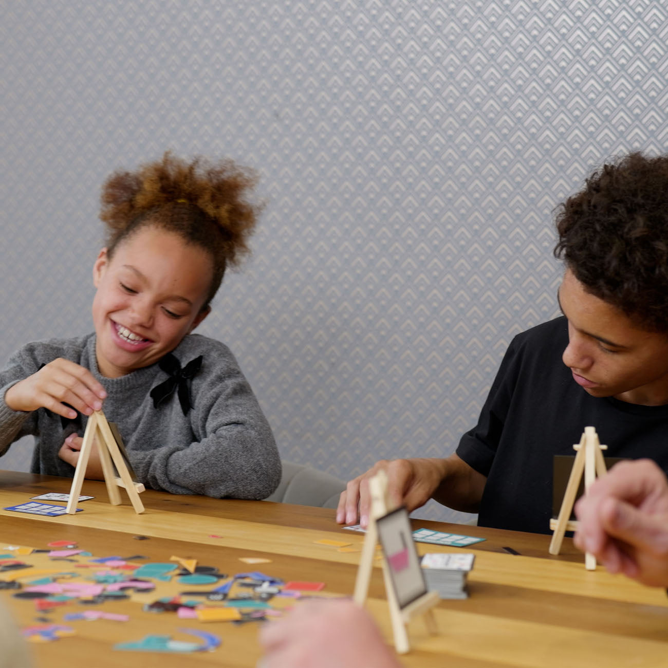 Two children sitting at a wooden table, smiling and playing with small wooden easels and colourful game pieces against a grey patterned wallpaper background.