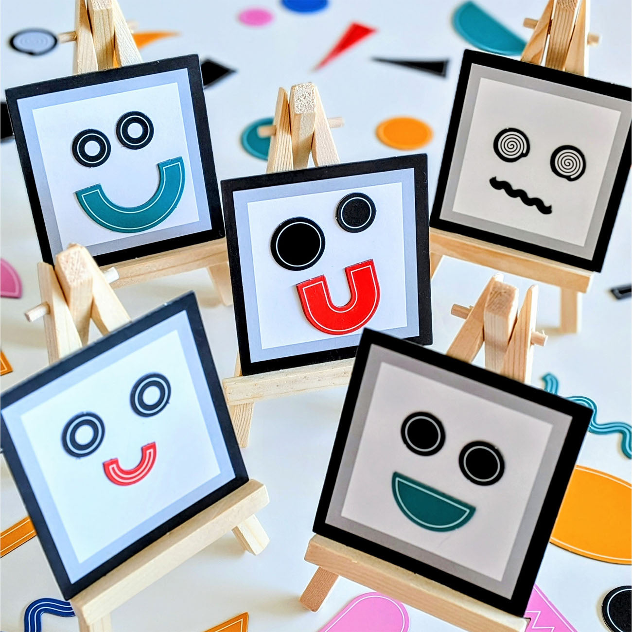 Five small framed art pieces on wooden easels, each with a simple face made of black circular eyes and coloured curved mouth or nose shapes in teal, red, and wavy black lines.
