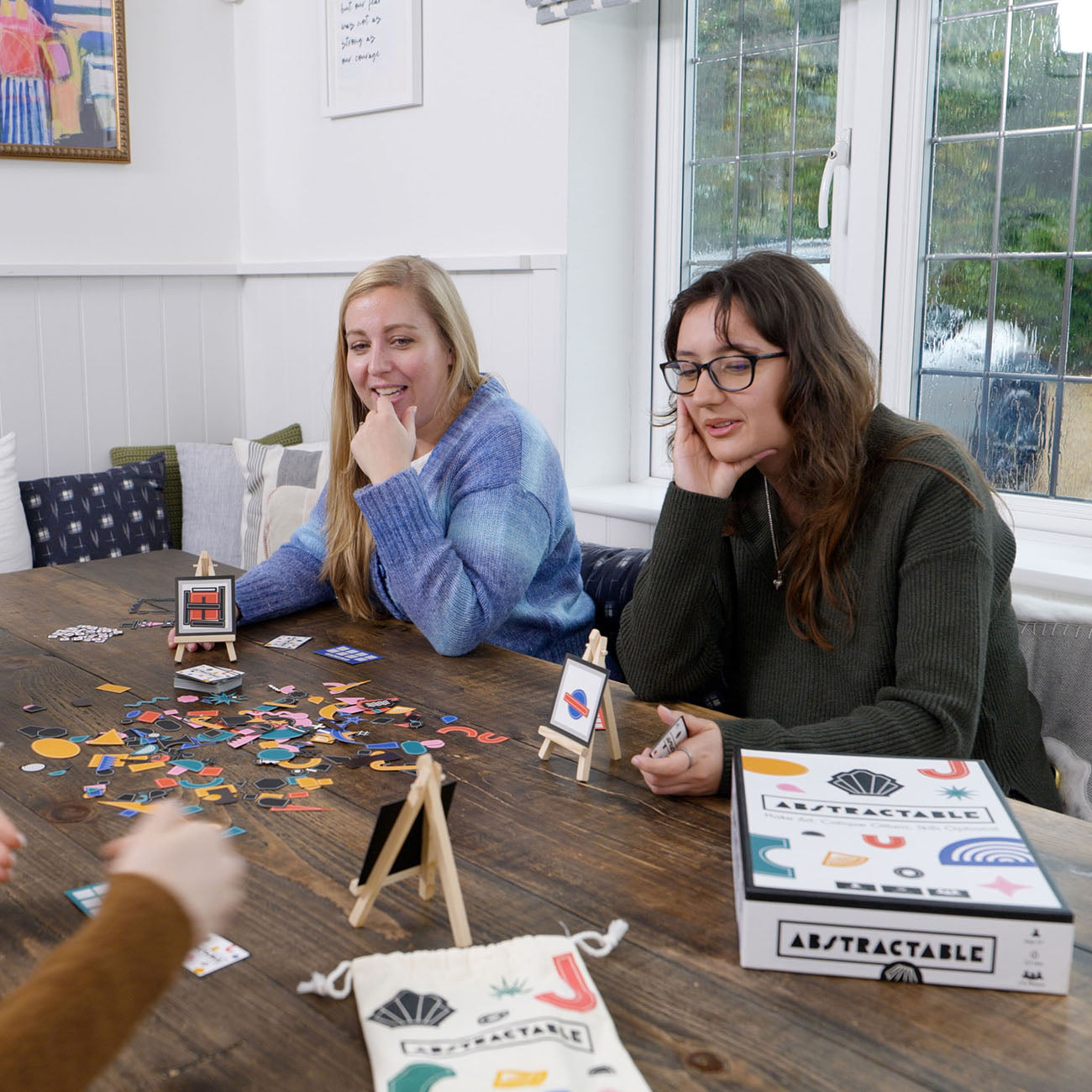 Two women sitting at a wooden table playing a game with small canvas art prints and colourful game pieces. A box labelled 'ABSTRACTABLE' is visible on the table, along with a matching drawstring pouch and a small easel stand.