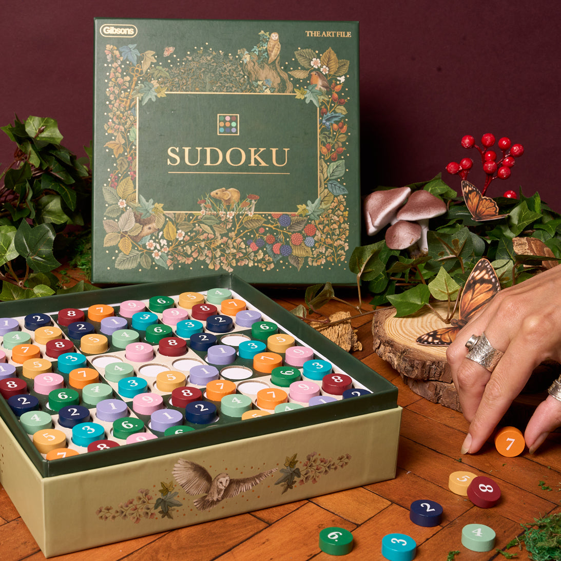 Sudoku game set with colorful pegs on a wooden surface, surrounded by decorative plants and a butterfly.