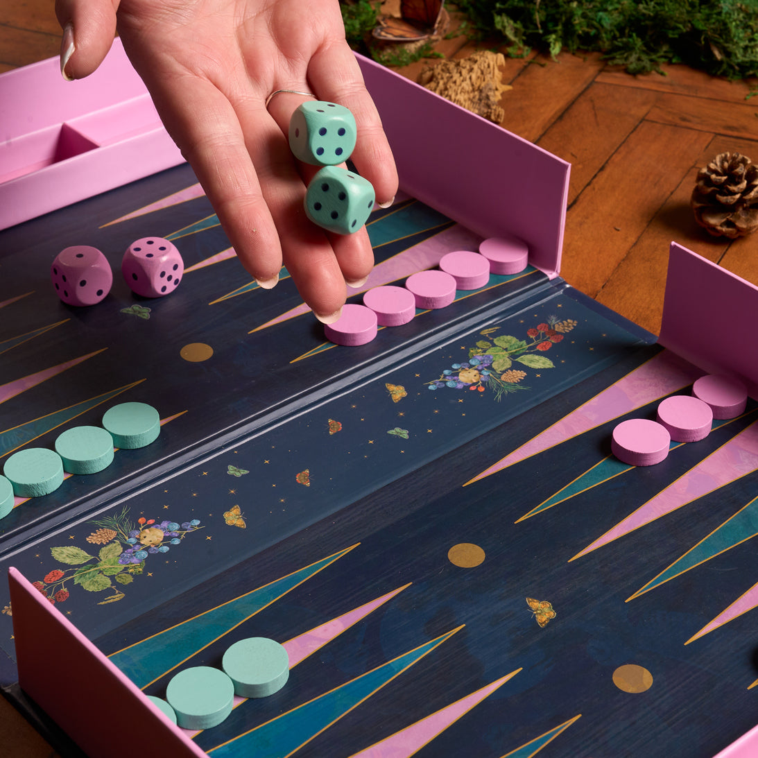 Hand holding dice over a decorative game board with pink and blue elements.
