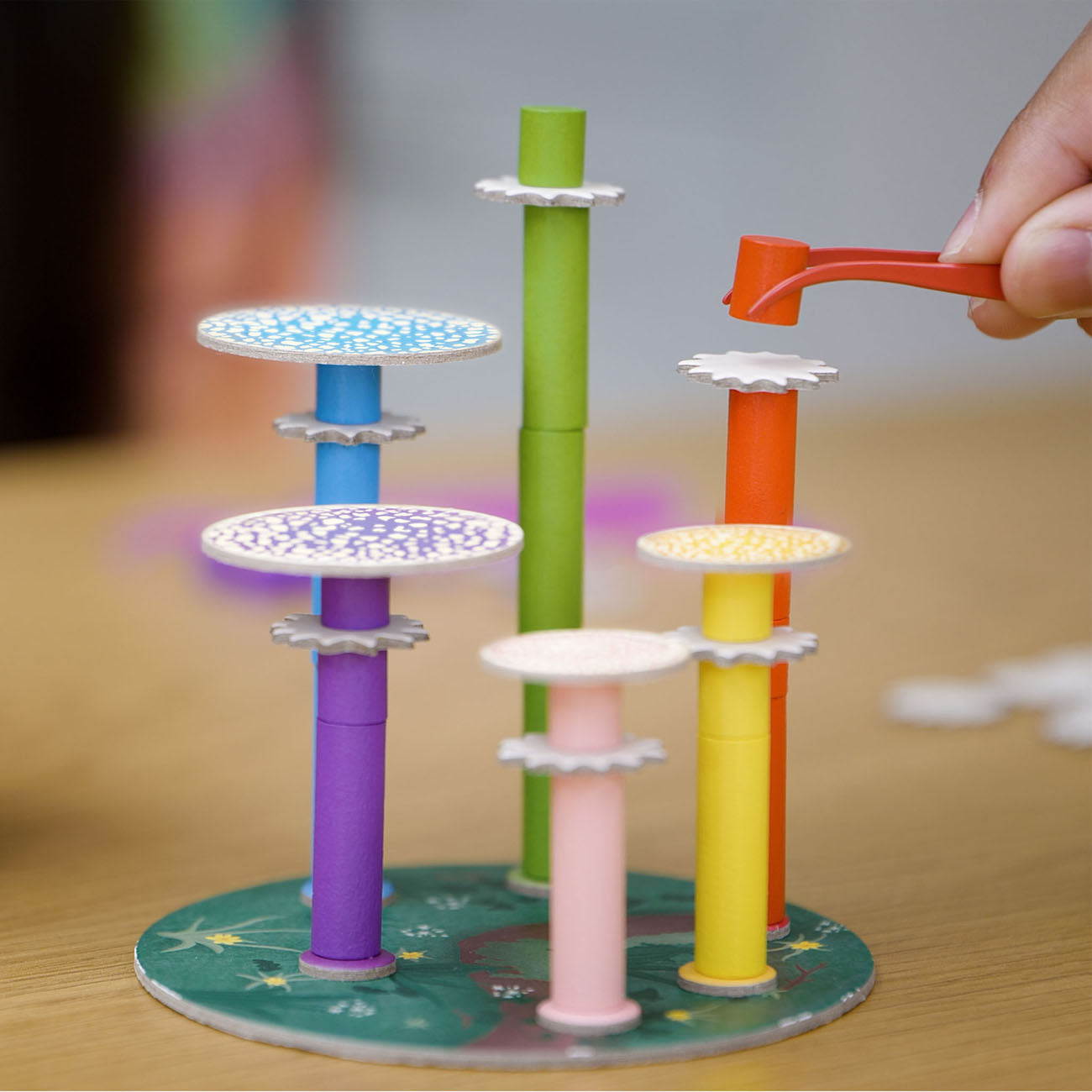 A colourful mushroom stacking toy set on a wooden tabletop, with a hand placing a small orange piece onto one of the mushroom caps.