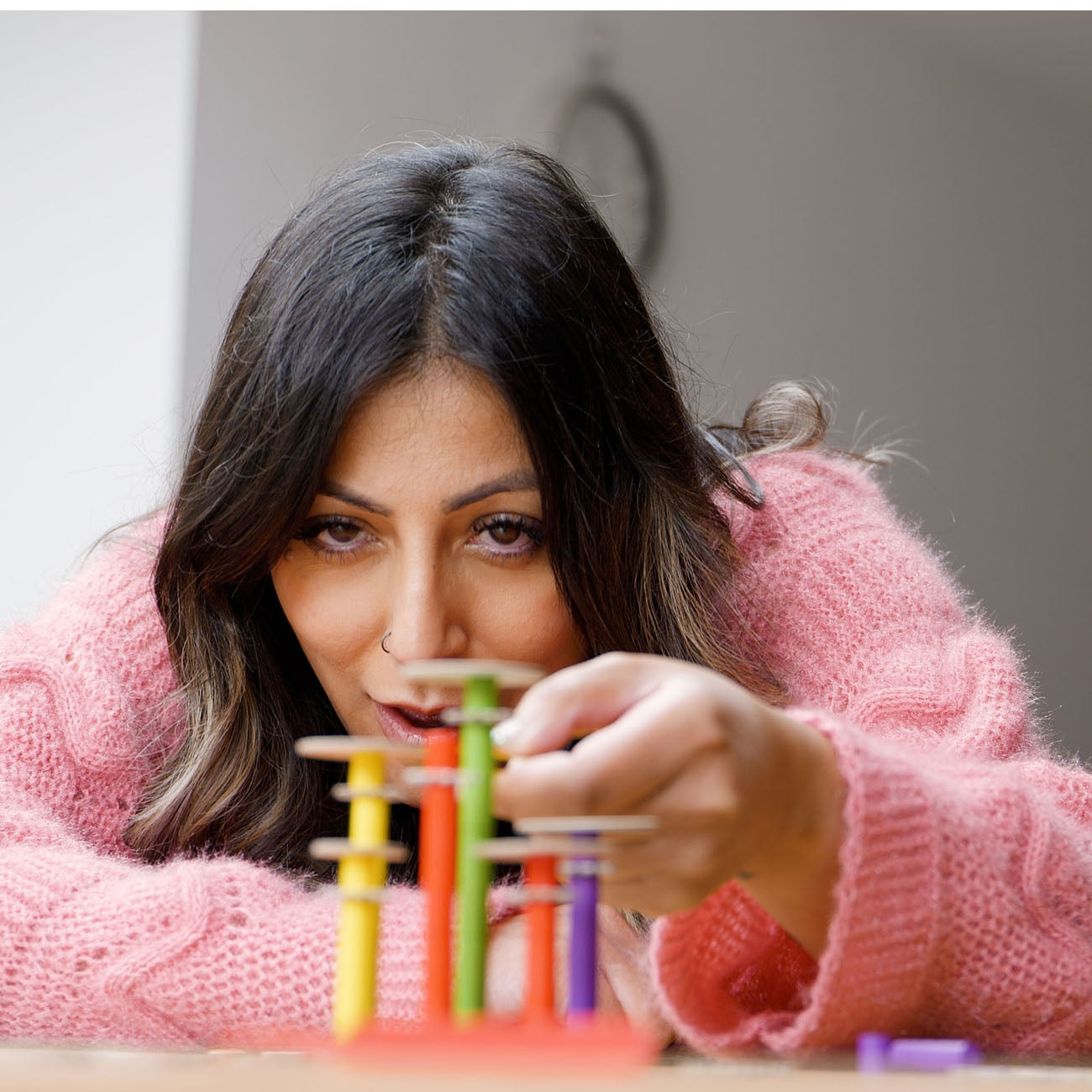 A woman with dark hair wearing a pink knit sweater leans over a colourful peg toy, focusing closely on it in a softly lit indoor setting.