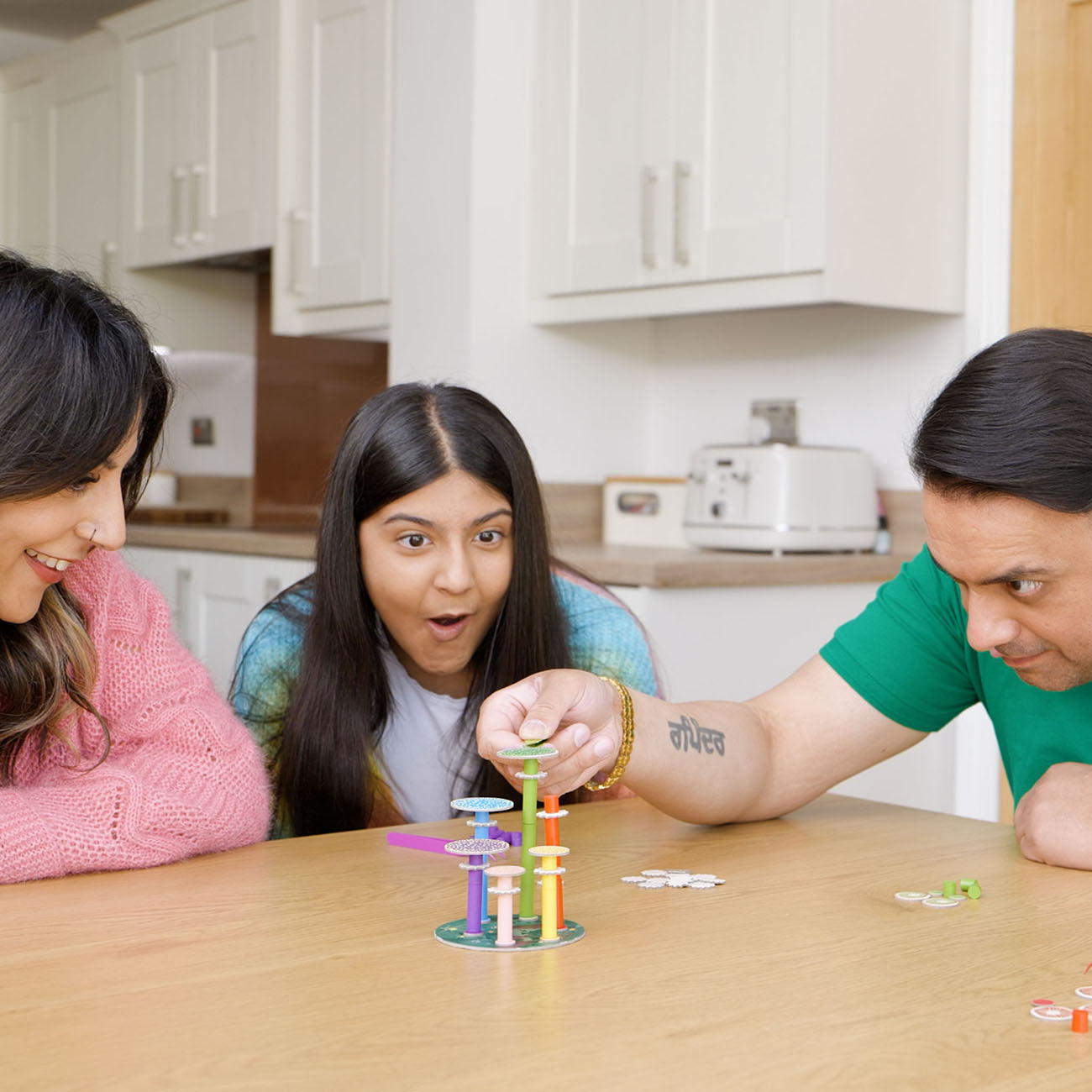 Three people gathered around a wooden table in a bright kitchen setting, playing with a colourful stacking game. The center person looks surprised, while the person on the right adjusts a tower of the toy.