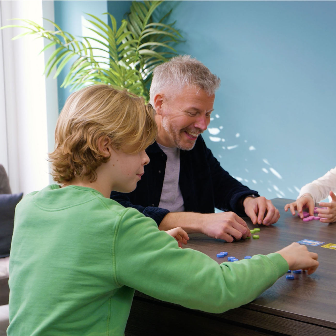 A man and a young boy are sitting at a wooden table playing a card game, with a woman's hand and another person's arm visible at the right edge. The room has a light blue wall and a green potted plant in the background.