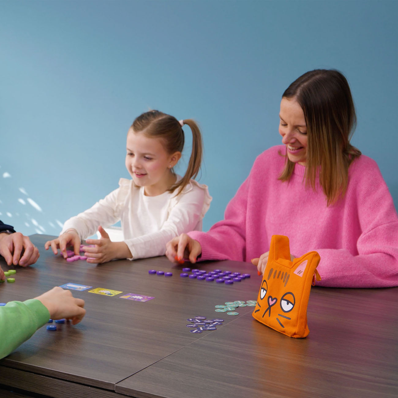 A woman and a young girl are playing a board game with purple tokens around a dark wooden table. An orange cat-shaped game pouch with a cartoon face sits on the table.