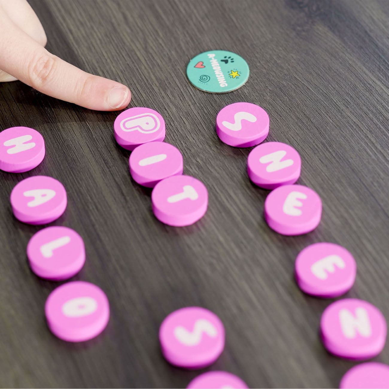 A close-up of a hand placing a pink circular letter tile with a white letter 'H' on a dark wood surface, alongside other matching pink letter tiles spelling out 'HELLO' and 'WORLD'. A small round teal sticker is visible near the top.