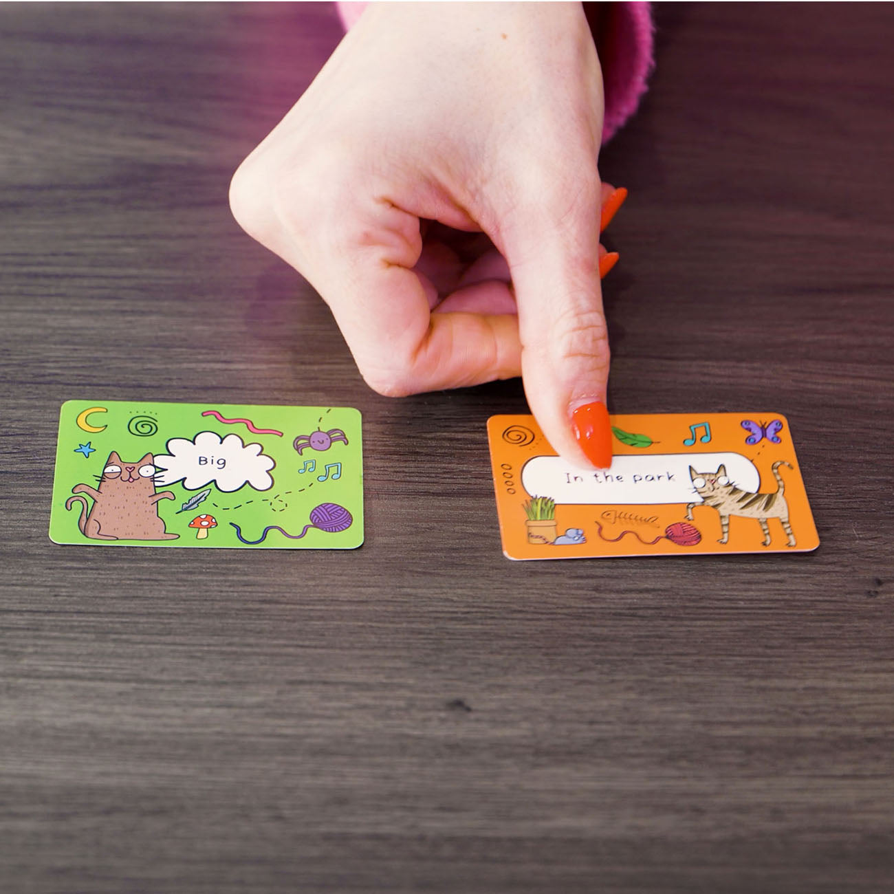 A close-up of a person's hand with orange nail polish pointing at a small illustrated card on a dark wooden table. Another card lies nearby on the table.