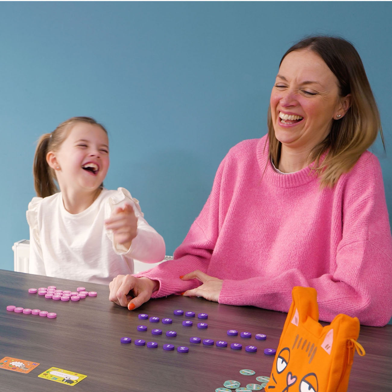 A woman and a young girl laughing together at a wooden table, playing with colourful game pieces and an orange cat-themed drawstring pouch against a solid blue background.