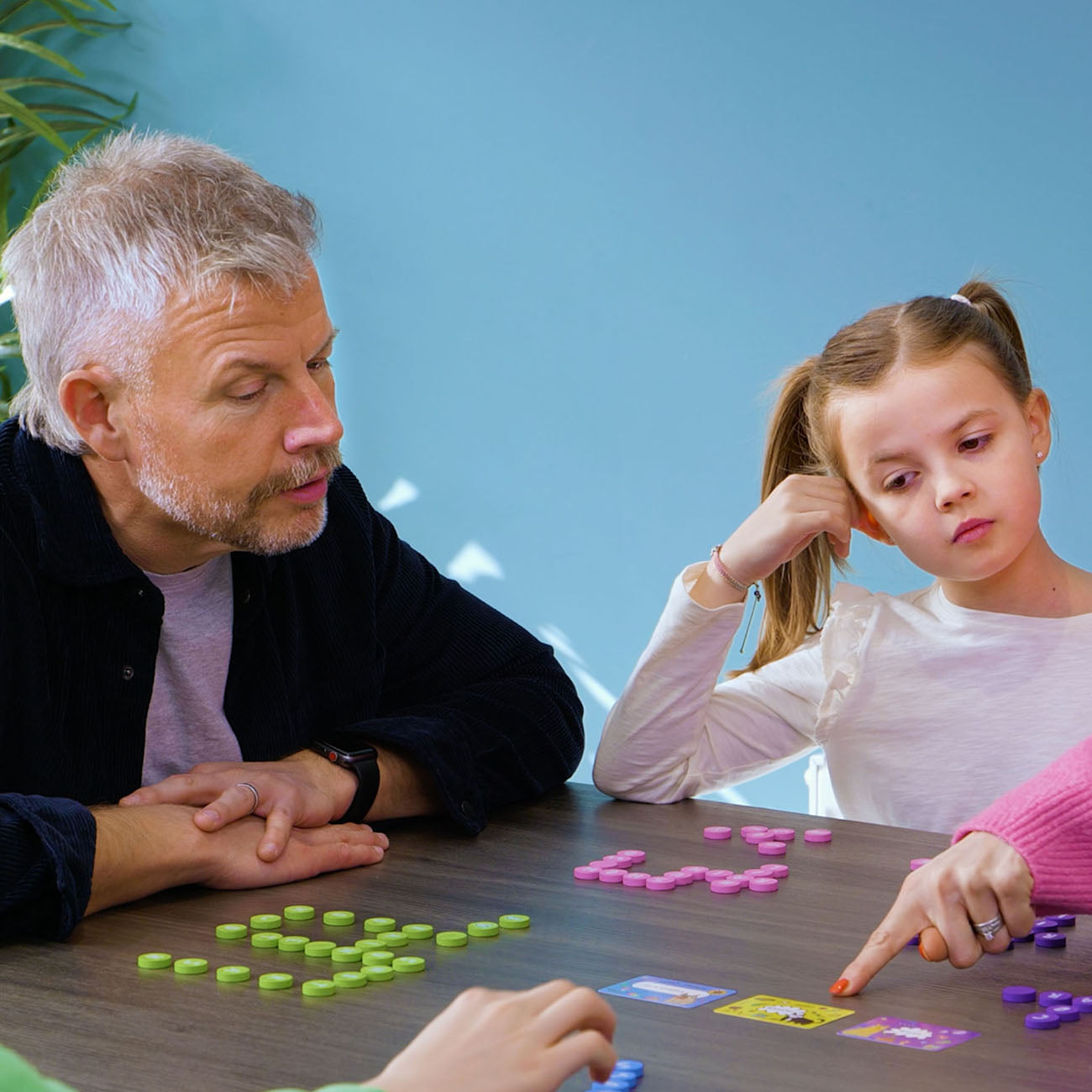 An adult man and a young girl are sitting at a wooden table playing a card game with colourful small tokens. The man is wearing a dark navy overshirt over a light purple t-shirt, and the girl is in a white long-sleeve top.