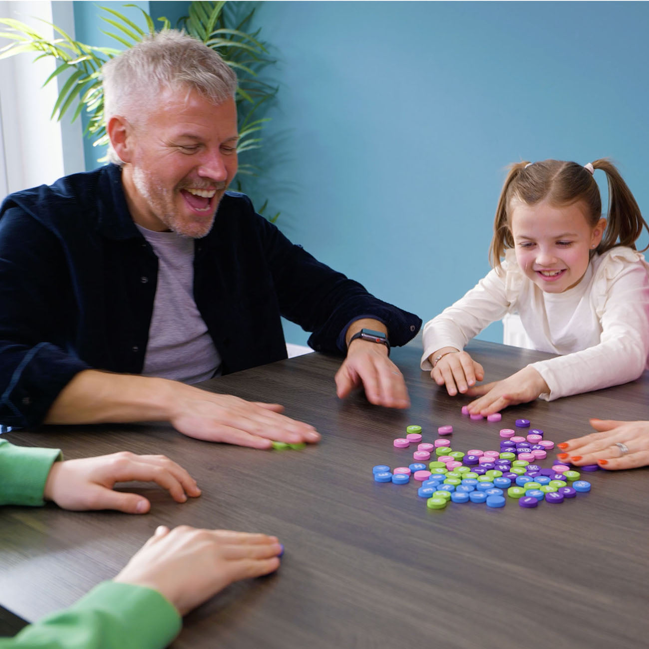 A man and a young girl are smiling and playing with colourful round game pieces on a dark wooden table, with other people's hands visible at the edges of the frame.