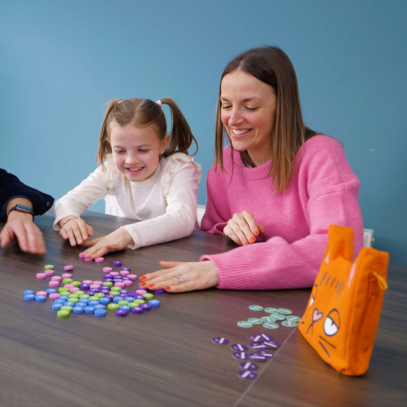 A woman and a young girl are sitting at a table playing with colorful round game tokens, with an orange character bag on the table. The woman is wearing a pink sweater, and the background is a solid blue wall.