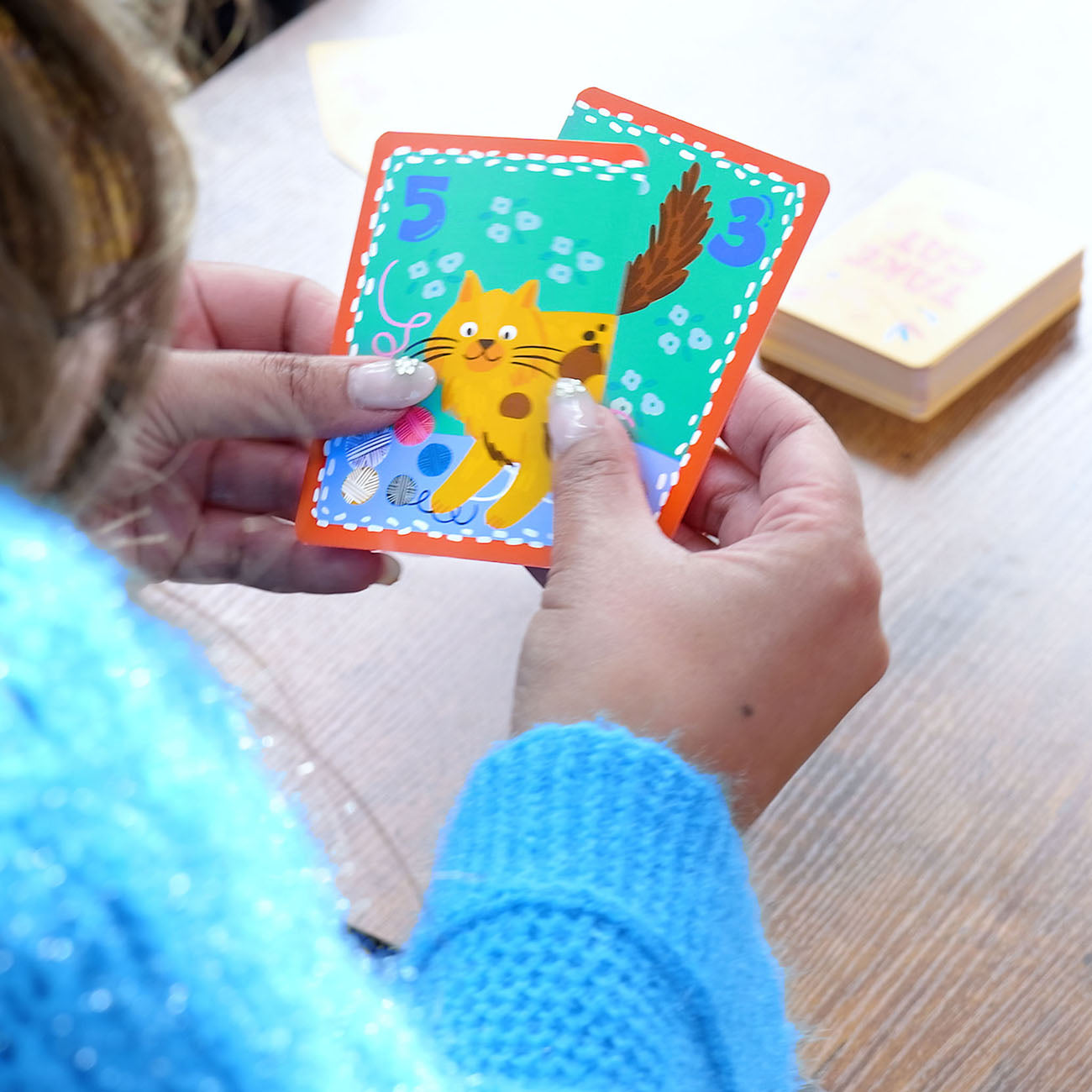 A person holding two colourful animal cards in both hands over a wooden table. The cards feature bright illustrations, including an orange cat on the front card with the number 5, and a brown tail on the back card with the number 3.